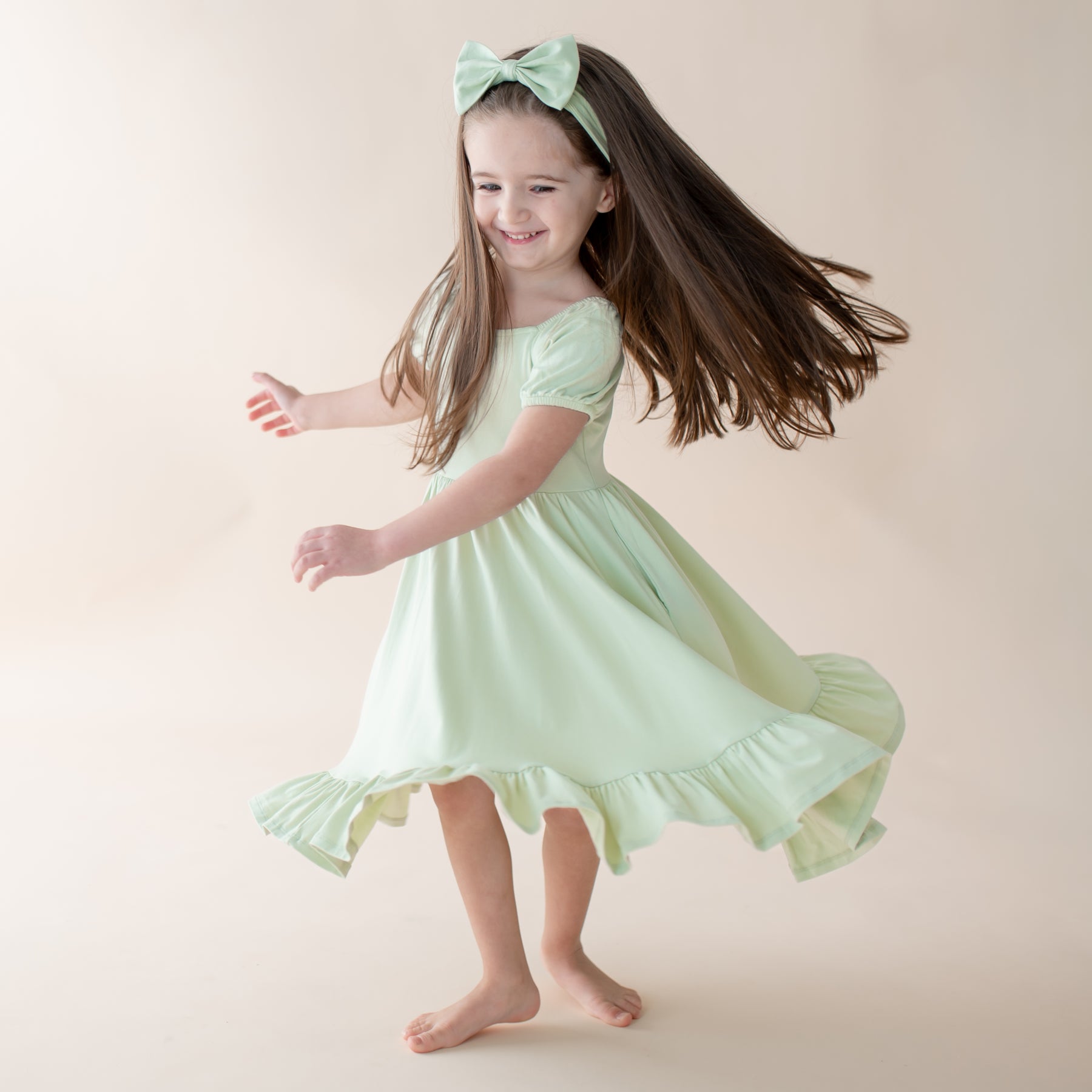Young girl in a light green dress with a bow in her hair, twirling against a plain background.