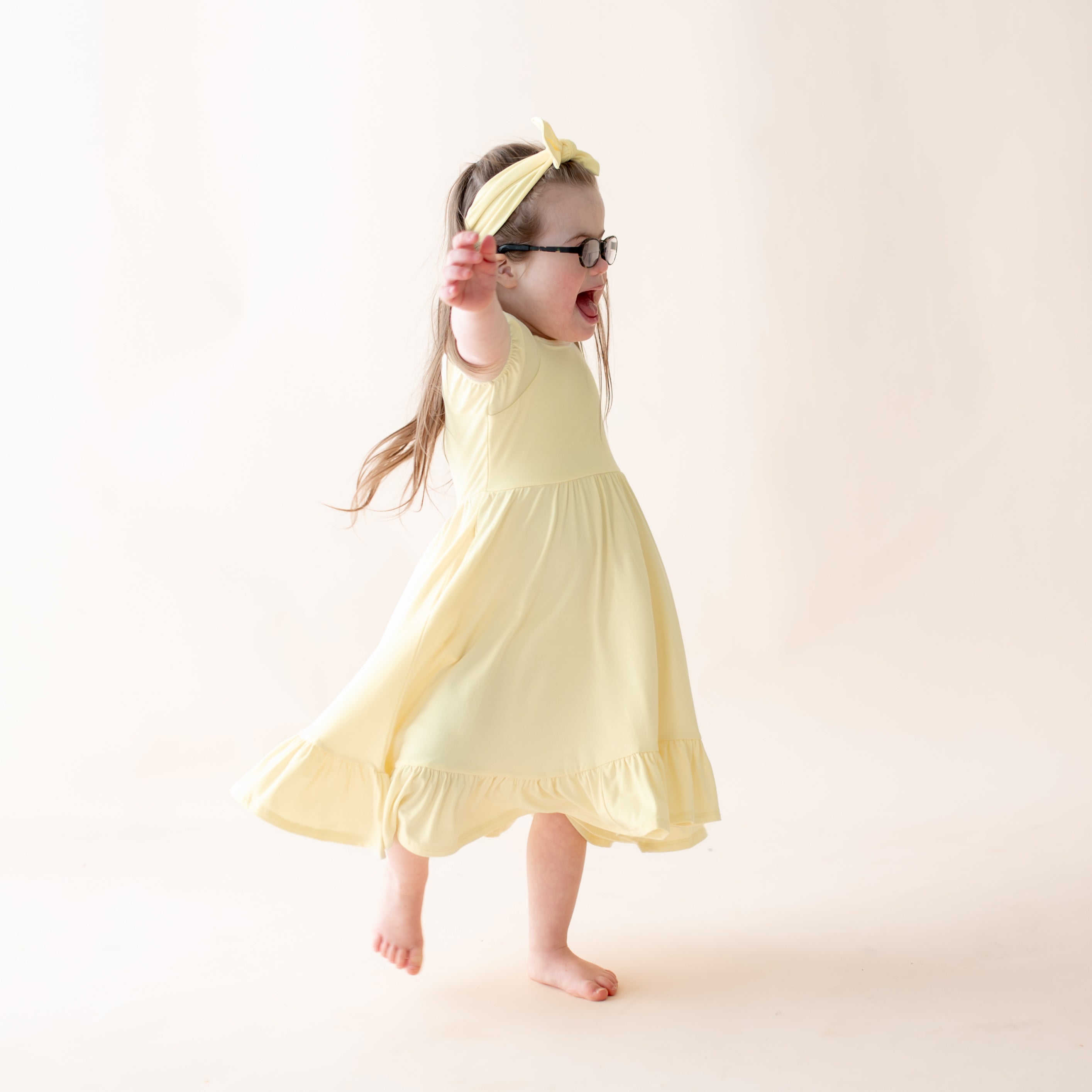 Child twirling, wearing a yellow dress and glasses on a plain background