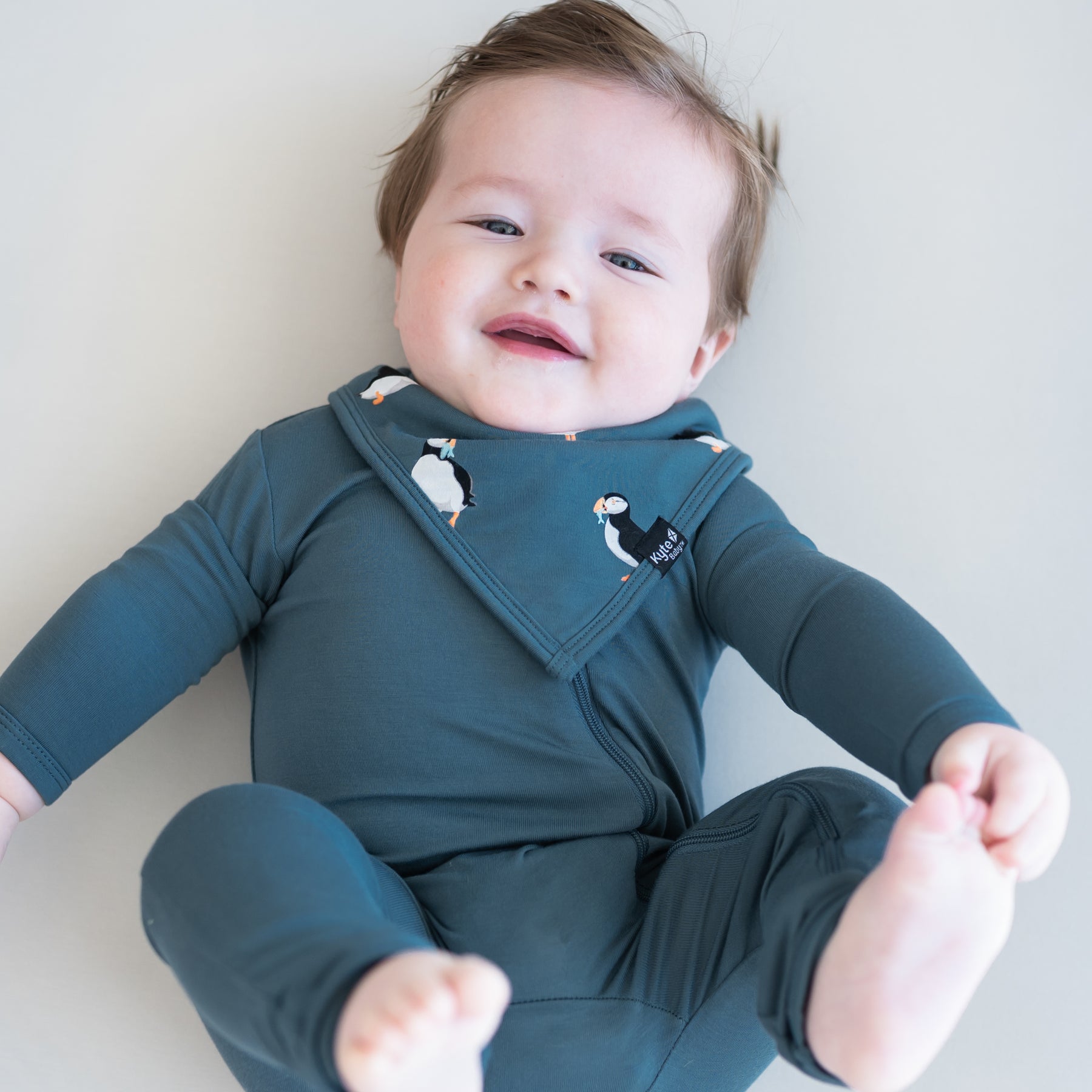Infant wearing the Bib in Puffin overtop an Atlantic Zippered romper laying on a light neutral surface