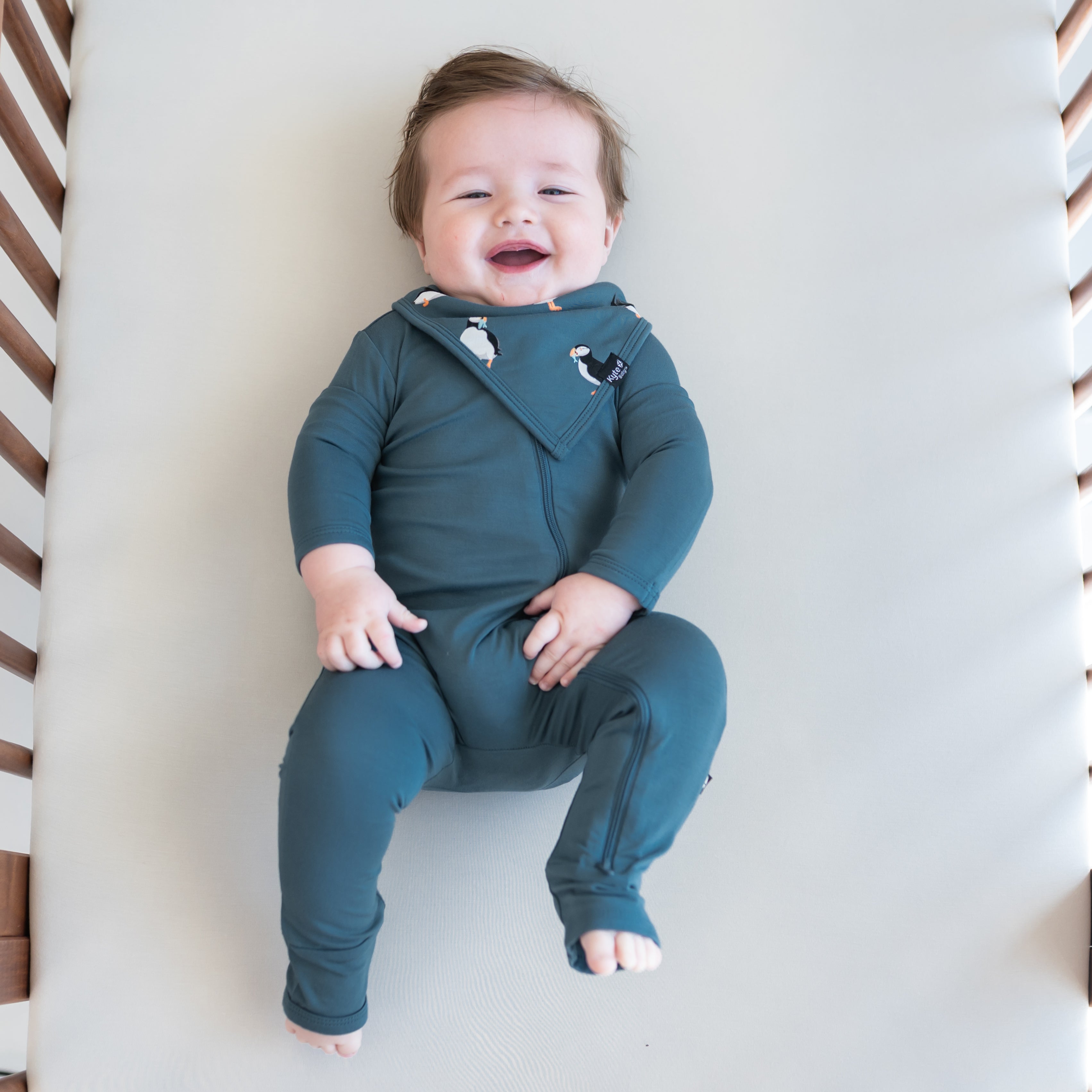 Infant laying in a crib wearing the Bib in Puffin and Atlantic zippered romper