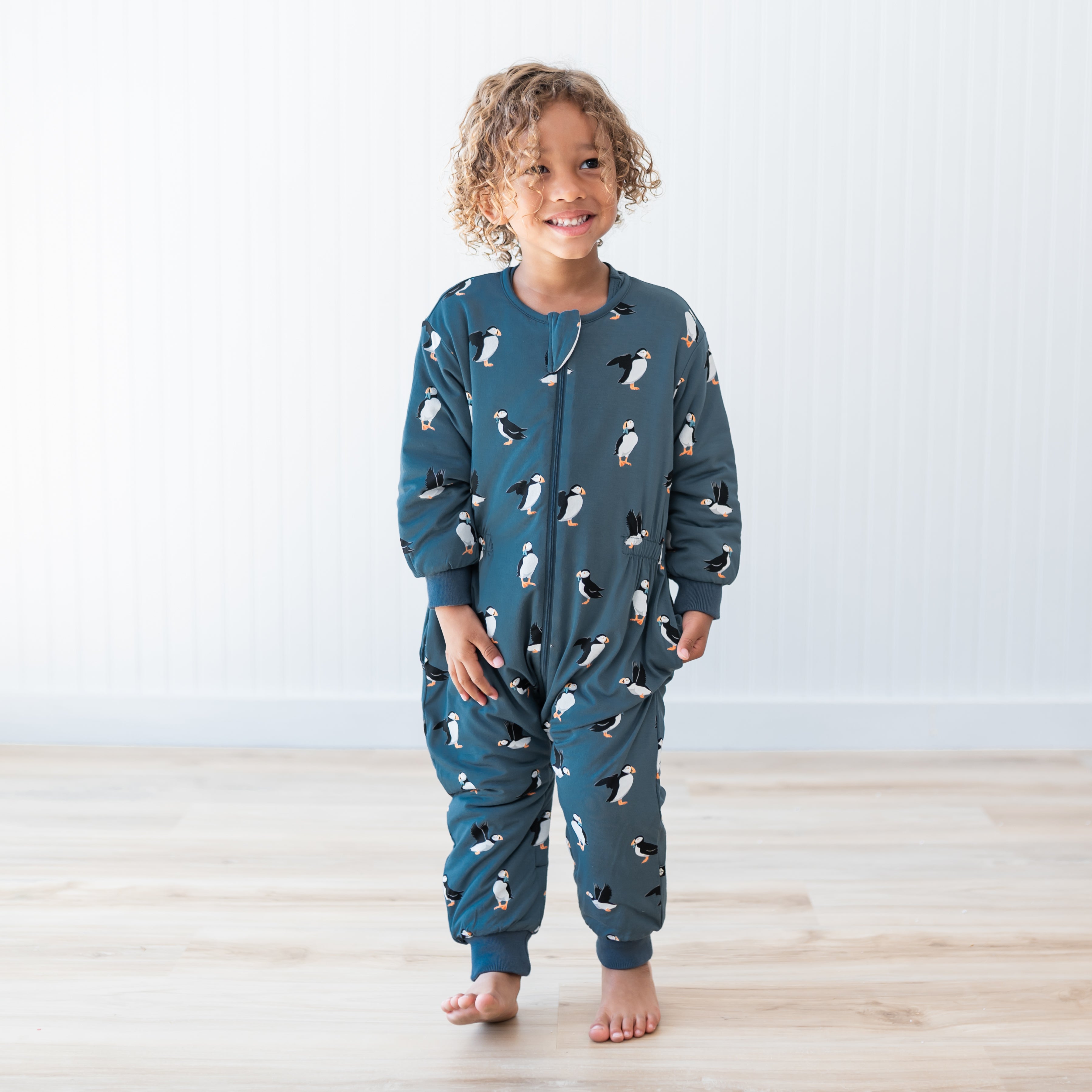 Young child standing on a light wood floor in front of a white paneled wall wearing the Cozy Playsuit in Puffin