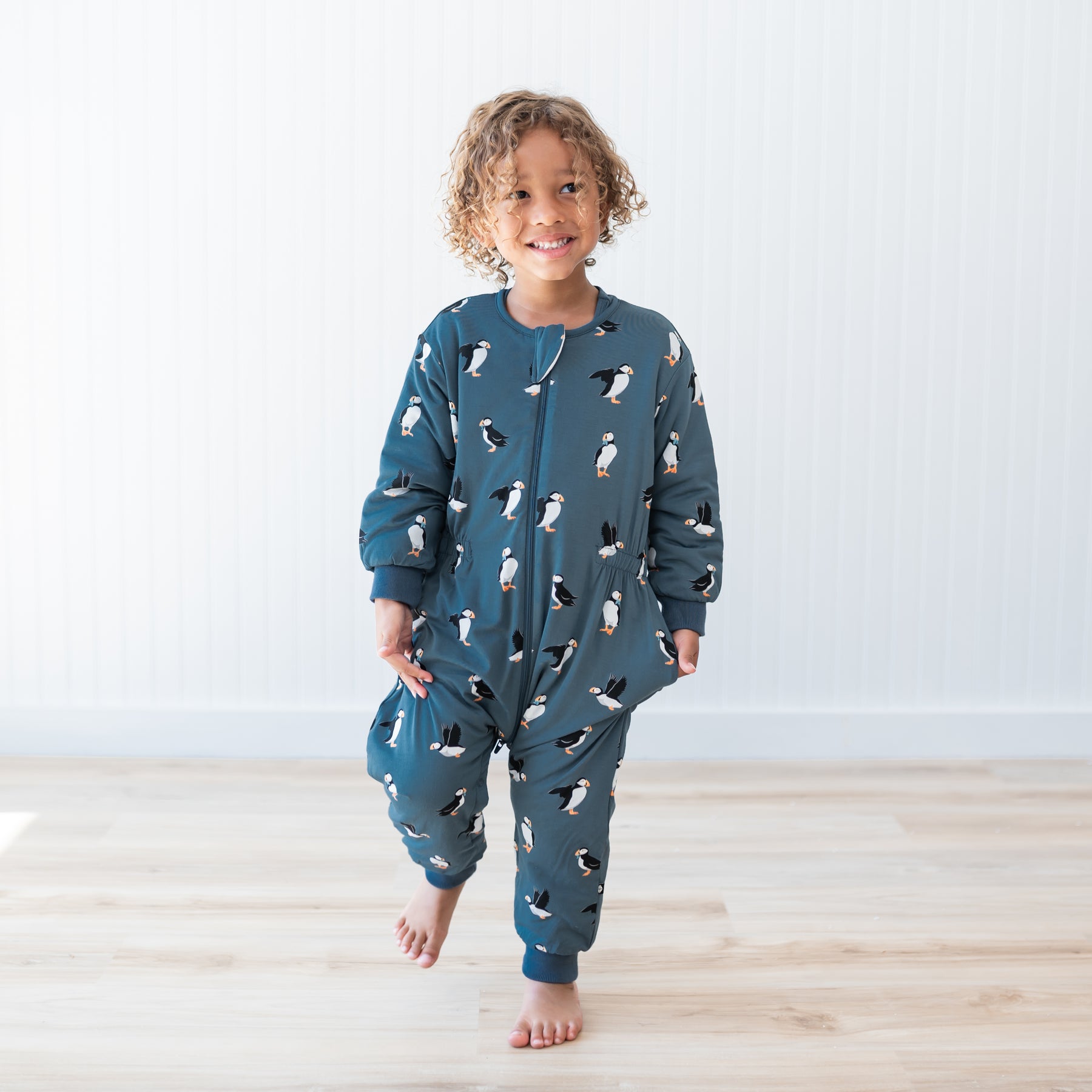 Young child walking on a light wood floor in front of a white paneled wall wearing the Cozy Playsuit in Puffin
