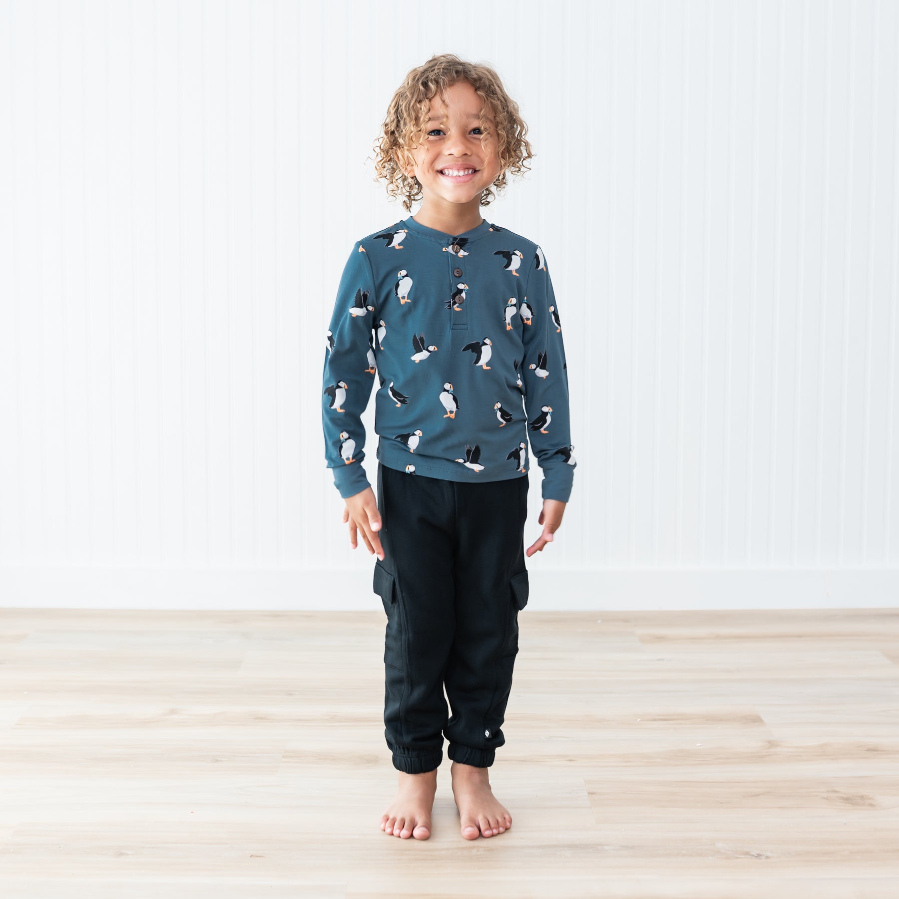 Smiling child standing on a light wood grain floor in front of a white paneled wall wearing the Long Sleeve Toddler Henley Tee in Puffin and black fleece cargo pants
