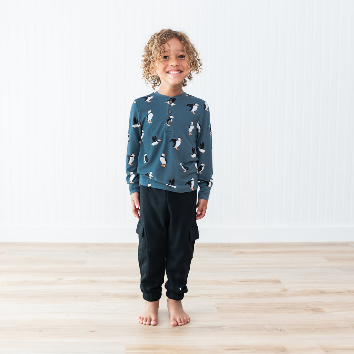 Young smiling child stand with their hands at their side wearing the Long Sleeve Toddler Henley Tee in Puffin with the black fleece cargo pants