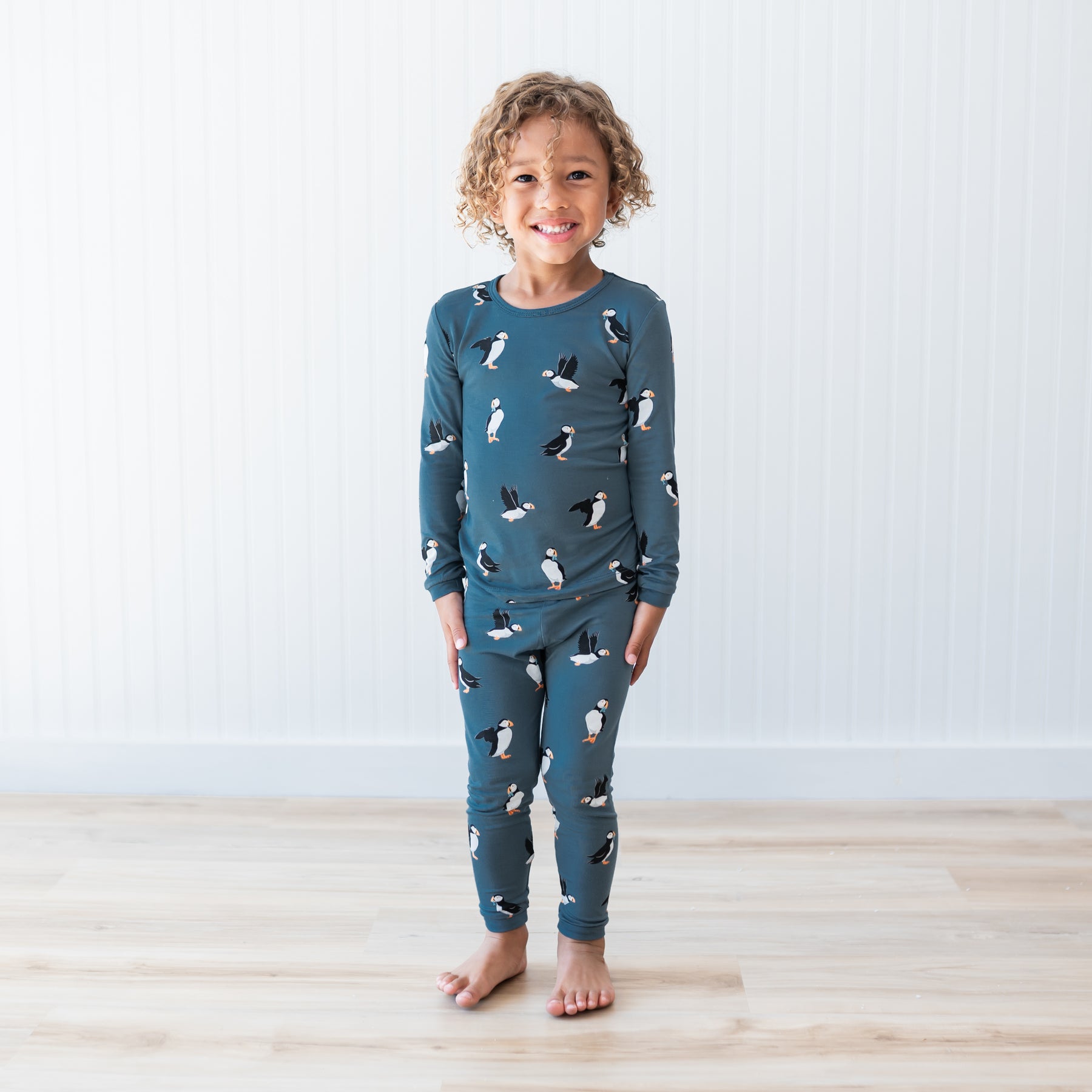 Young child standing with their hands at their sides in front of a white paneled wall wearing the Long Sleeve Pajamas in Puffin