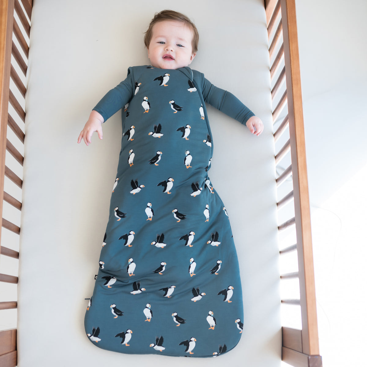 Young toddler laying in a crib wearing the Sleep Bag in Puffin 2.5 with an Atlantic long sleeve romper on underneath
