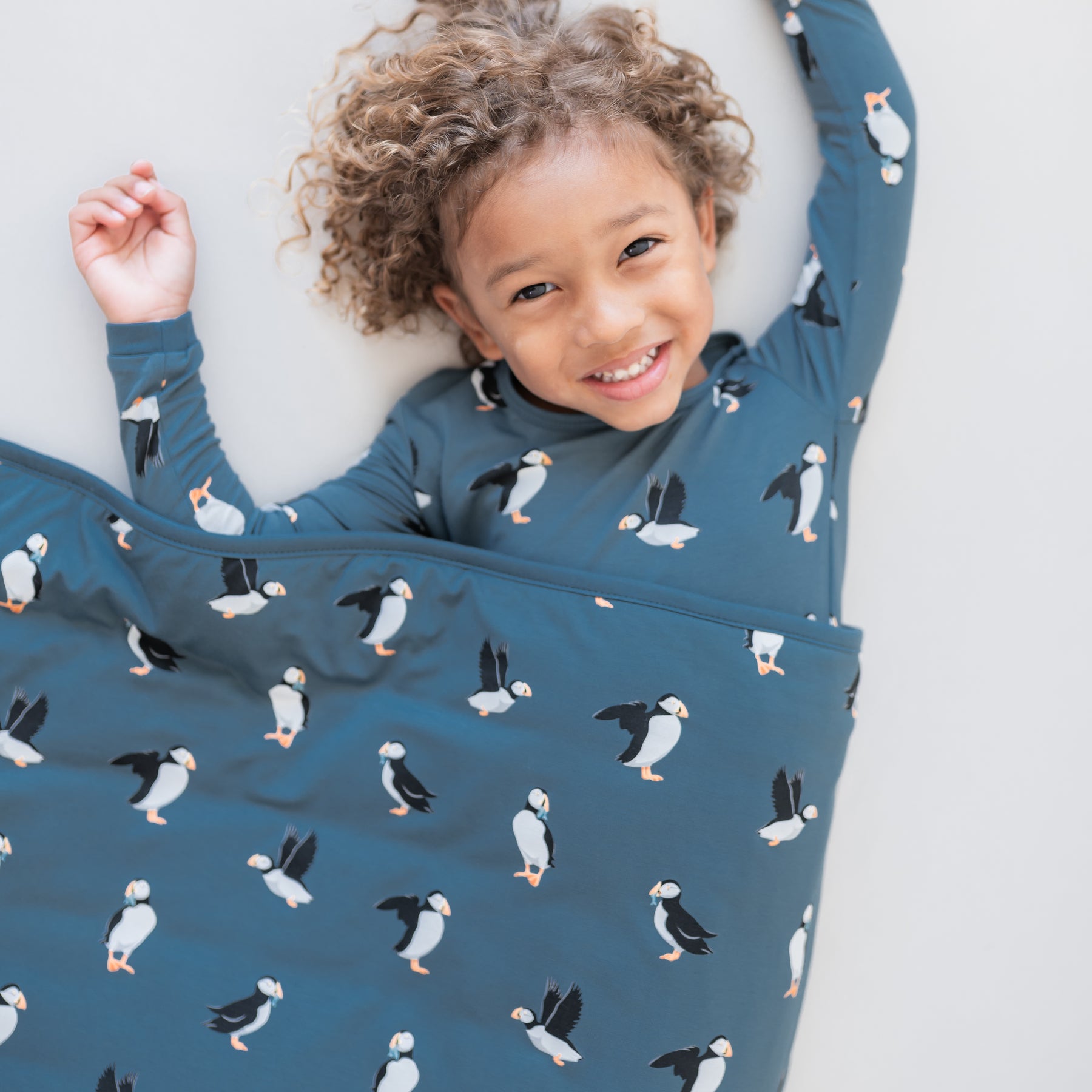 Young child laying down on a light neutral surface wearing the Long Sleeve Pajamas in Puffin with a matching toddler blanket overtop of them