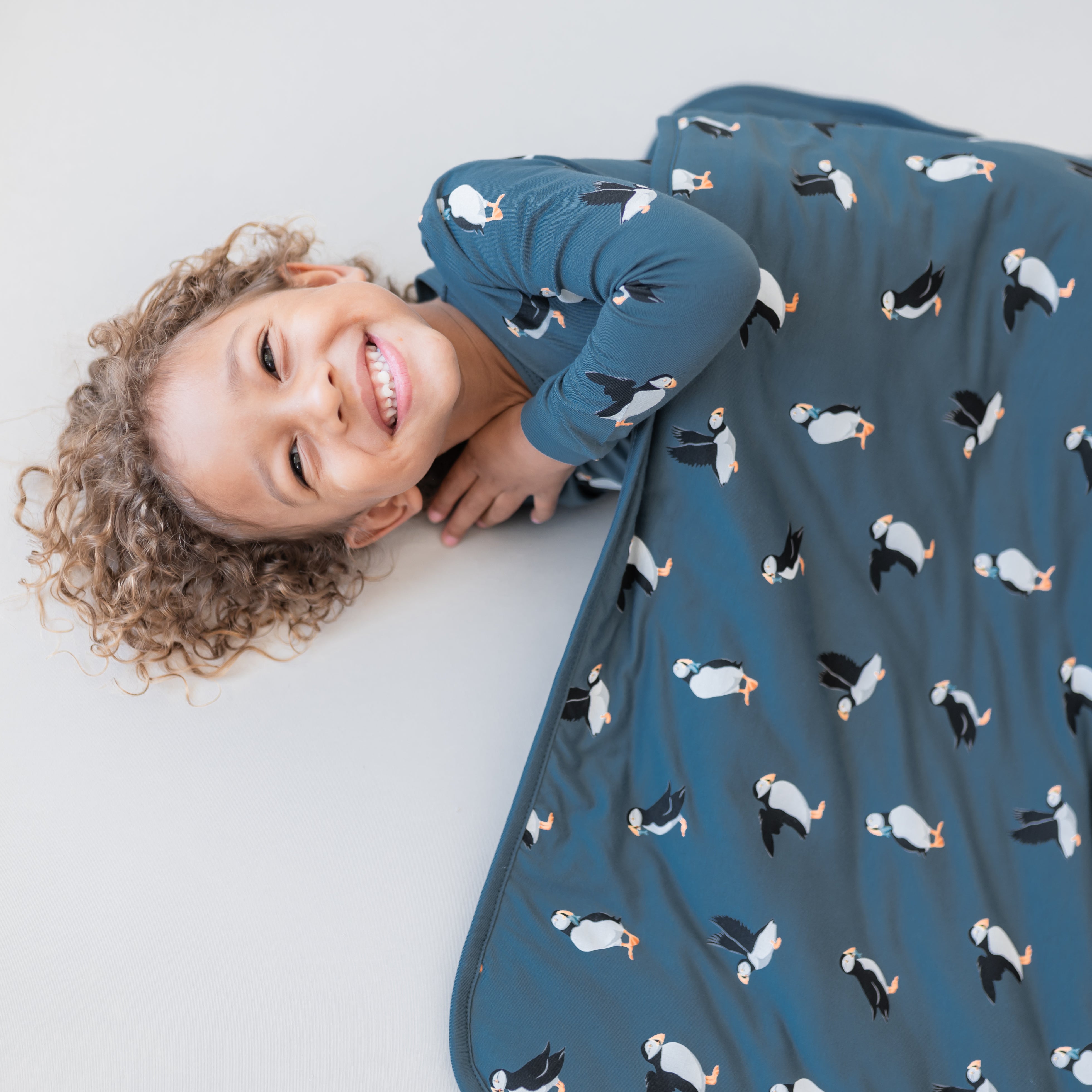 Smiling child laying down on a light neutral surface underneath a Toddler Blanket in Puffin 1.0 wearing matching long sleeve toddler pajamas