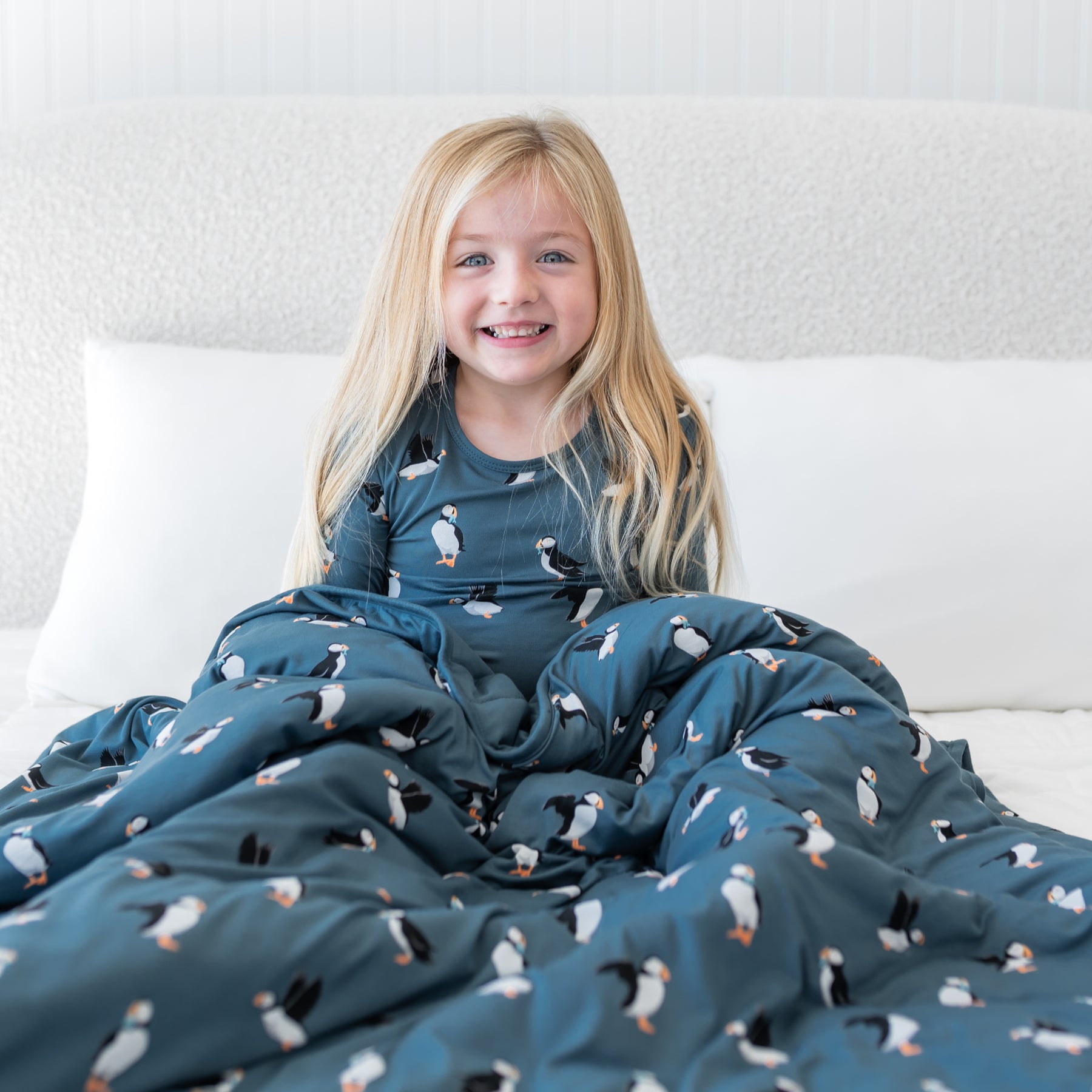 Young smiling girl sitting on a bed with the Youth Blanket in Puffin 2.5 over her lap wearing matching long sleeve toddler pajamas
