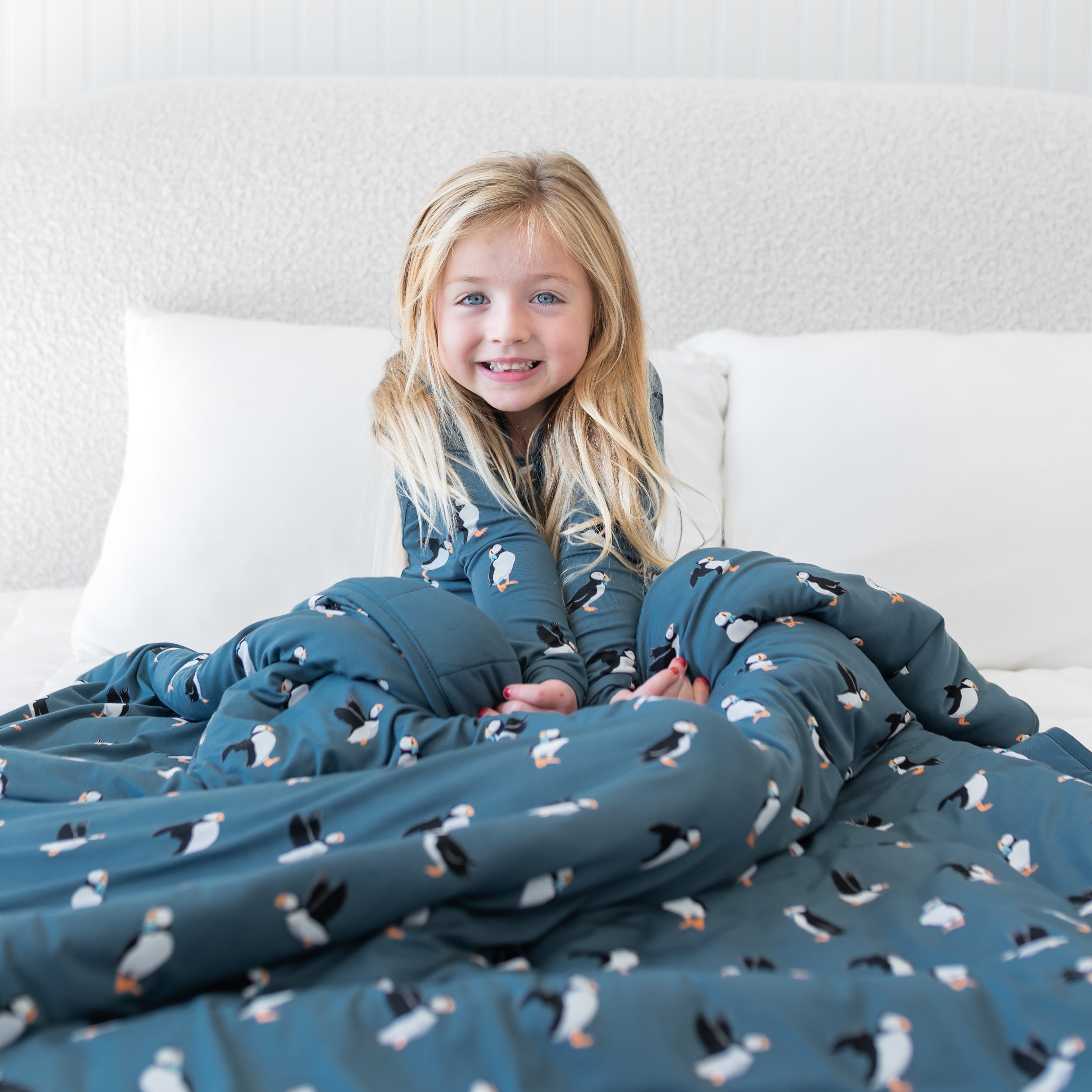 Smiling girl sitting in bed with the Youth Blanket in Puffin 2.5 overtop of her legs wearing matching long sleeve toddler pajamas