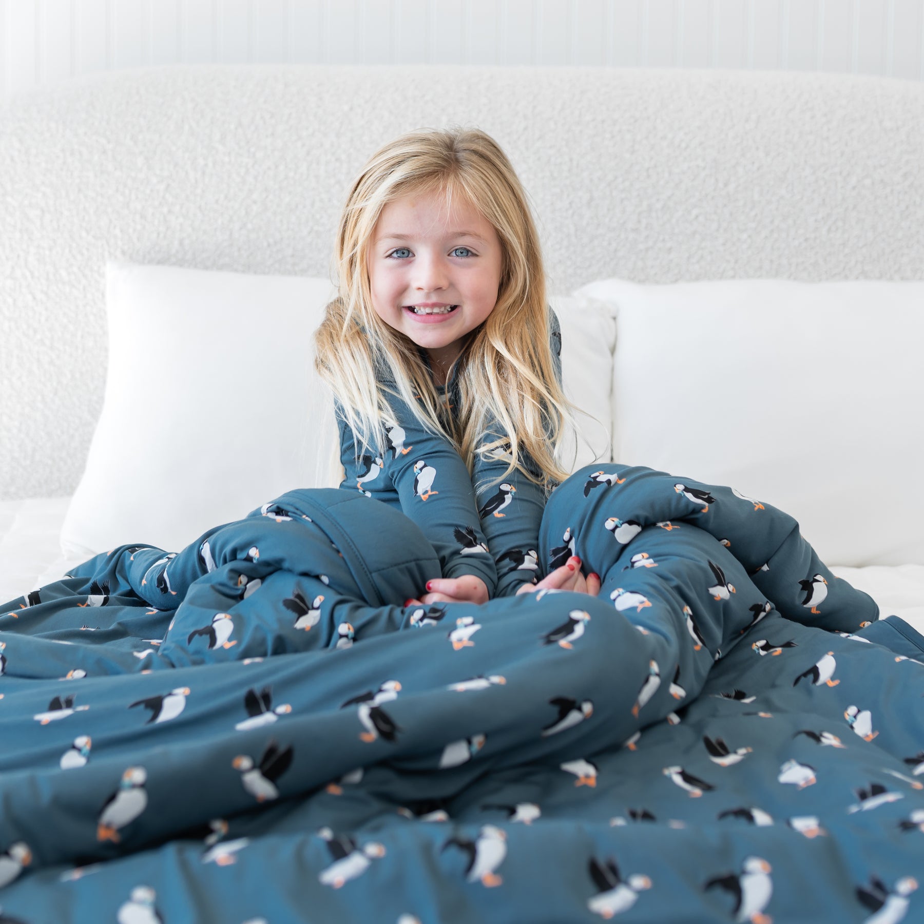 Smiling girl sitting in bed with the Youth Blanket in Puffin 2.5 overtop of her legs wearing matching long sleeve toddler pajamas