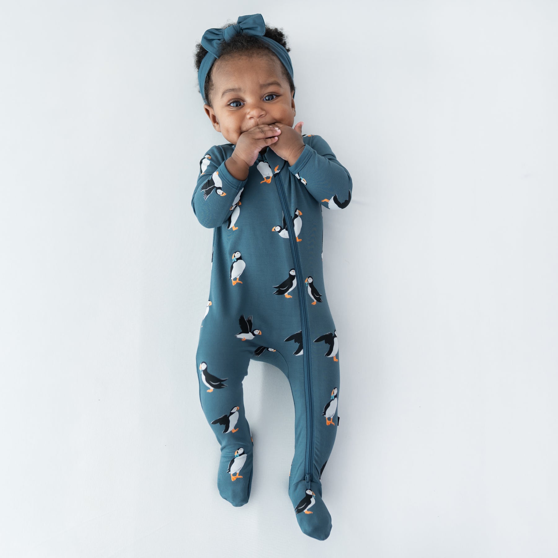 Young infant girl laying on a light colored surface sucking her fingers wearing the Zippered Footie in Puffin with a knotted bow headband in Atlantic
