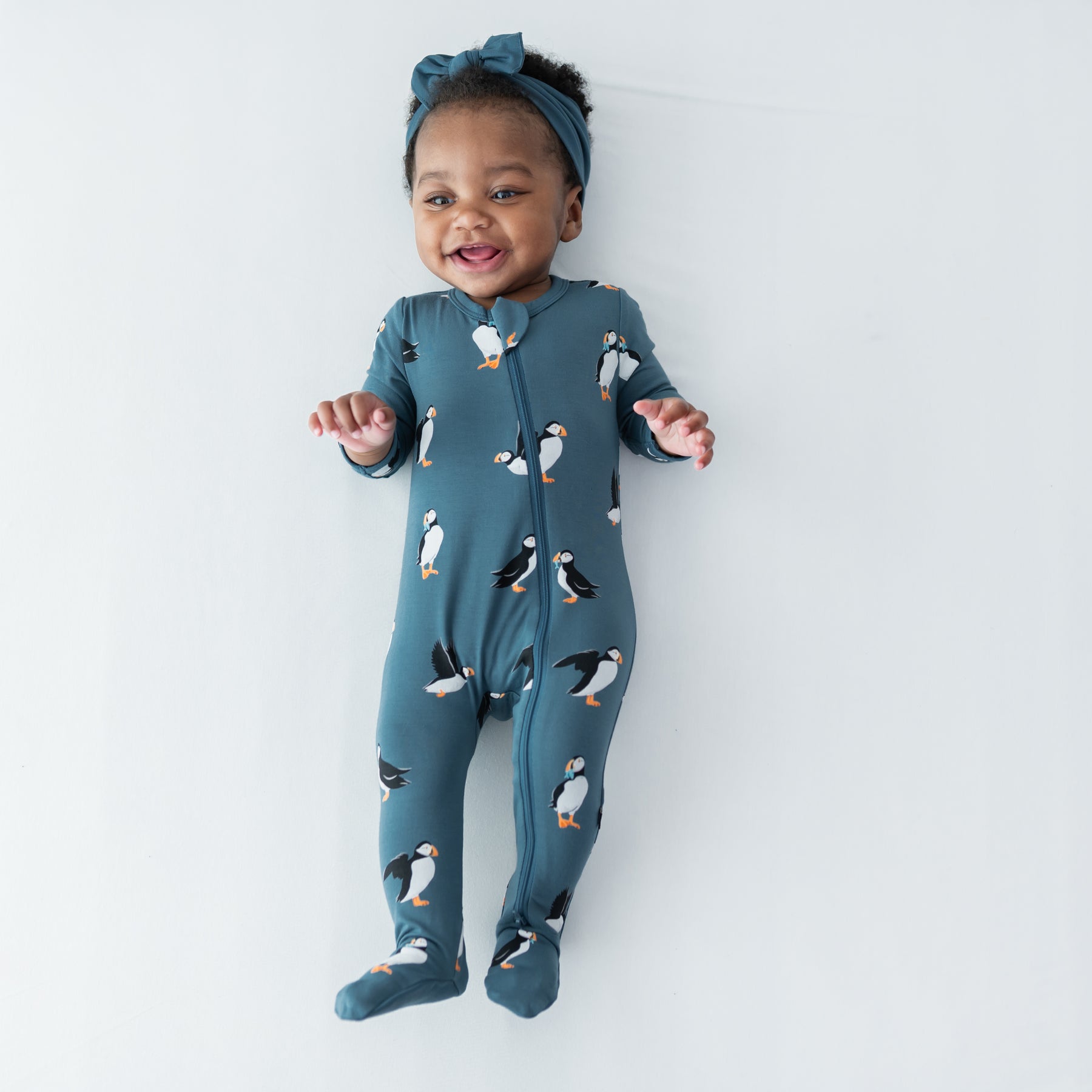 Young infant girl laying on a light colored surface wearing the Zippered Footie in Puffin with a knotted bow headband in atlantic