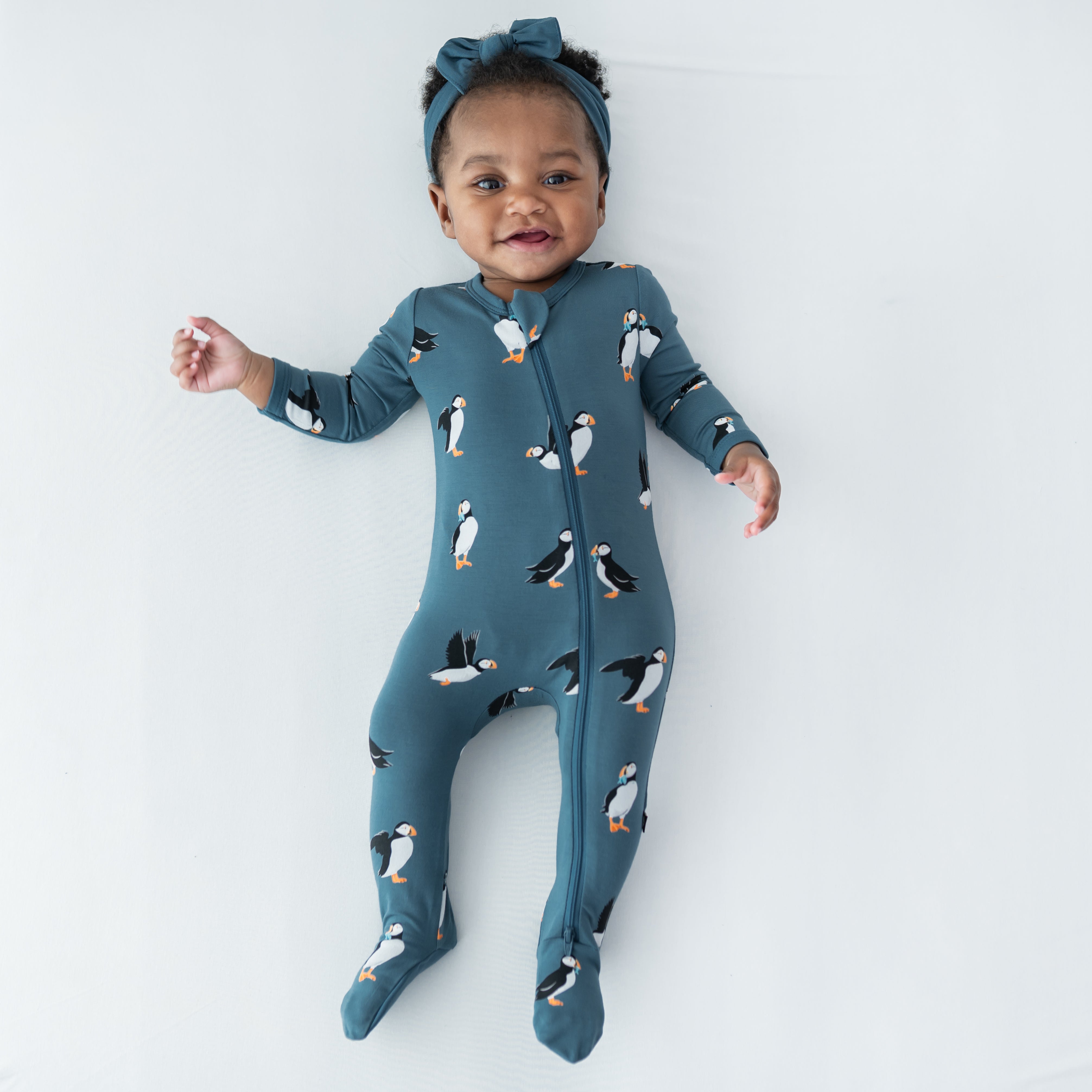 Young infant laying on a light neutral surface wearing the Zippered Footie in Puffin with a knotted bow headband in atlantic