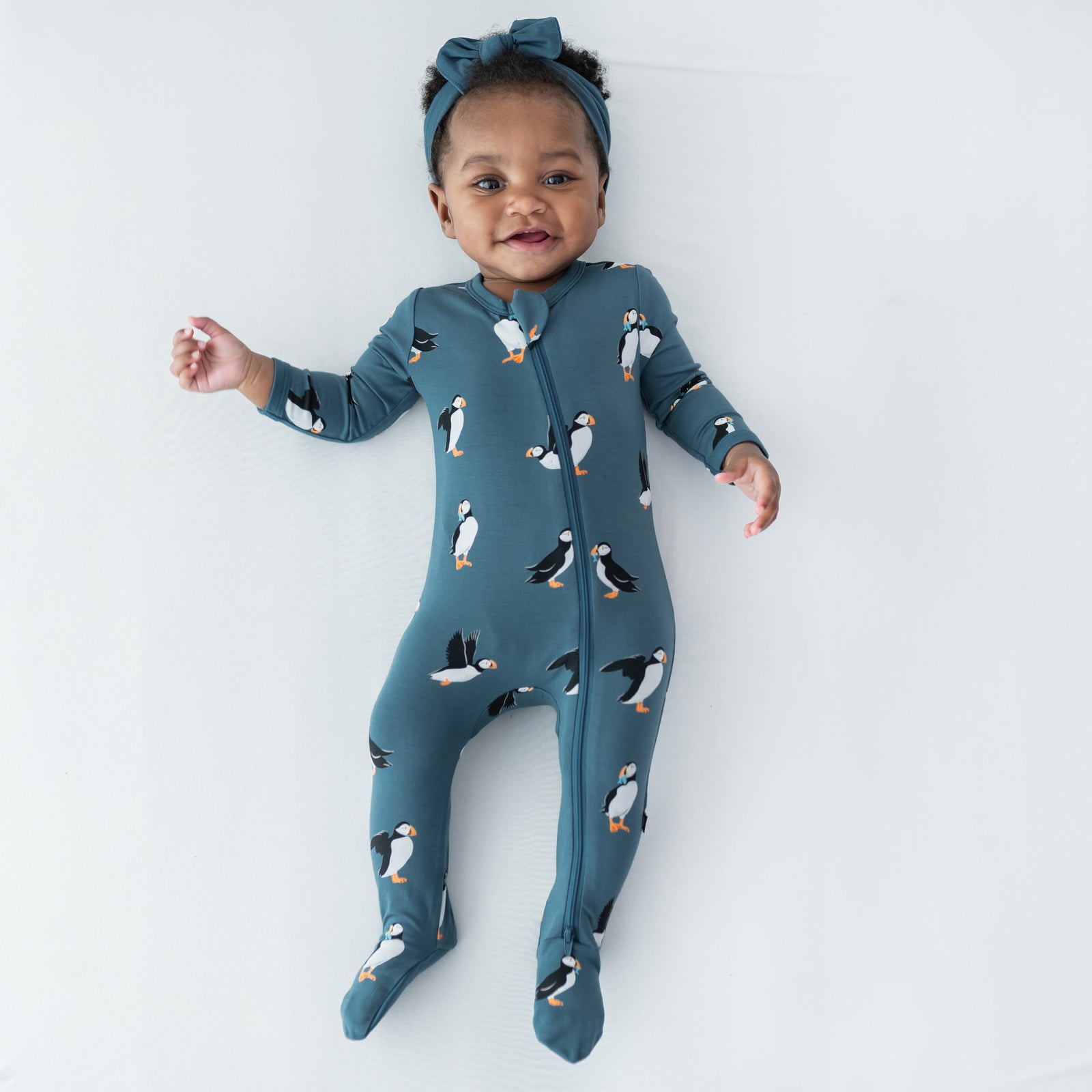 Young infant laying on a light neutral surface wearing the Zippered Footie in Puffin with a knotted bow headband in atlantic