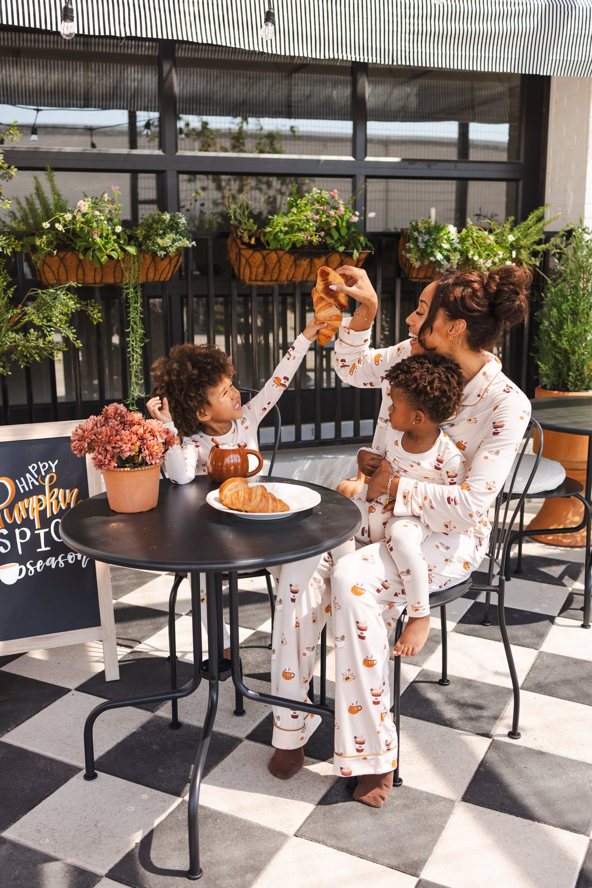 A family of three wearing matching white pajamas with a small pumpkin latte print, gathered around a small black table on a patio. The scene features a black and white checkered floor, potted plants, and a cozy breakfast setup.
