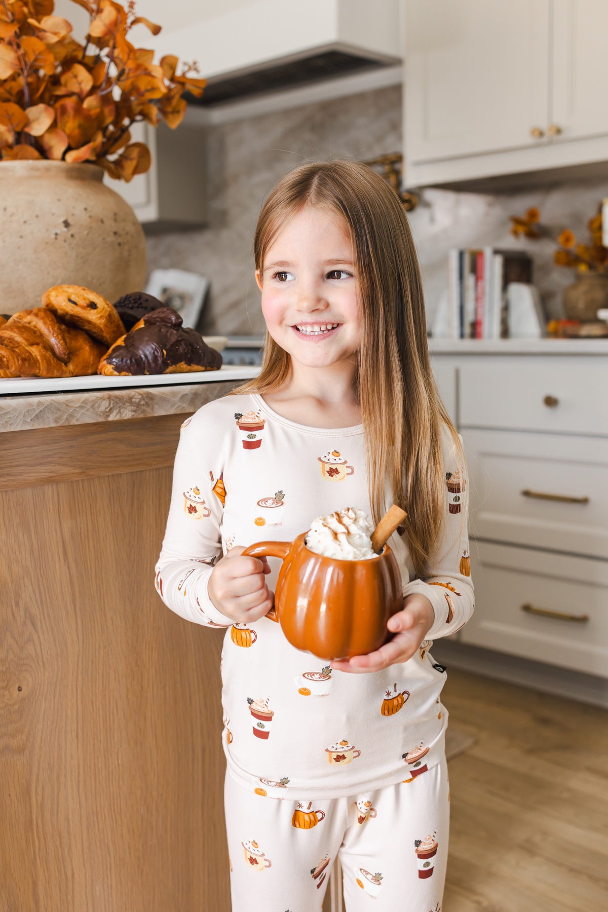 Girl smiling in kitchen wearing Long Sleeve Pajamas with Pumpkin Spice Latte print holding a spice latte cup.