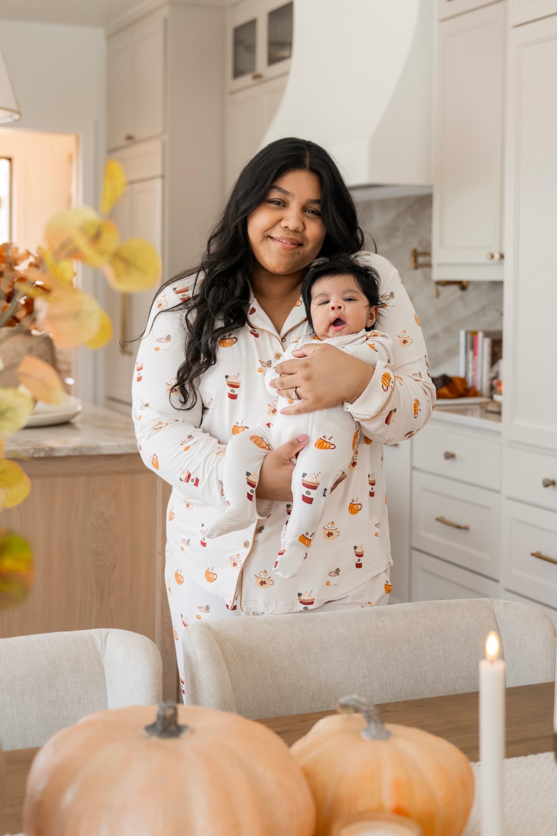 A woman in a kitchen holds a baby wearing matching white long-sleeve pajamas with a small orange and red pumpkin spice latte print. The scene includes pumpkins on a table in the foreground and white cabinets in the background.