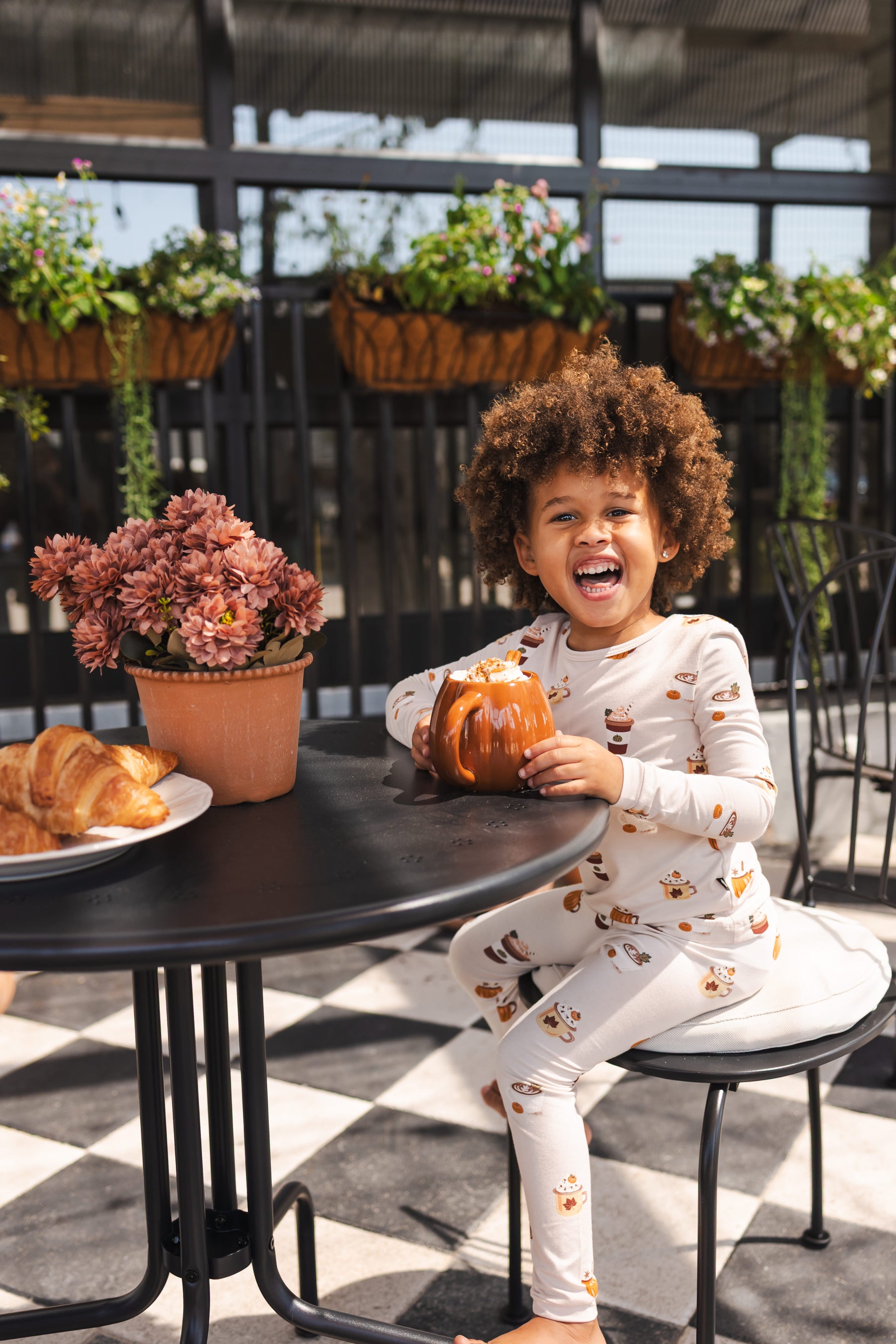 A young child sits smiling at an outdoor patio table, wearing a white long-sleeve pajama set with a brown coffee cup and mug pattern. The child is holding a small pumpkin decoration.