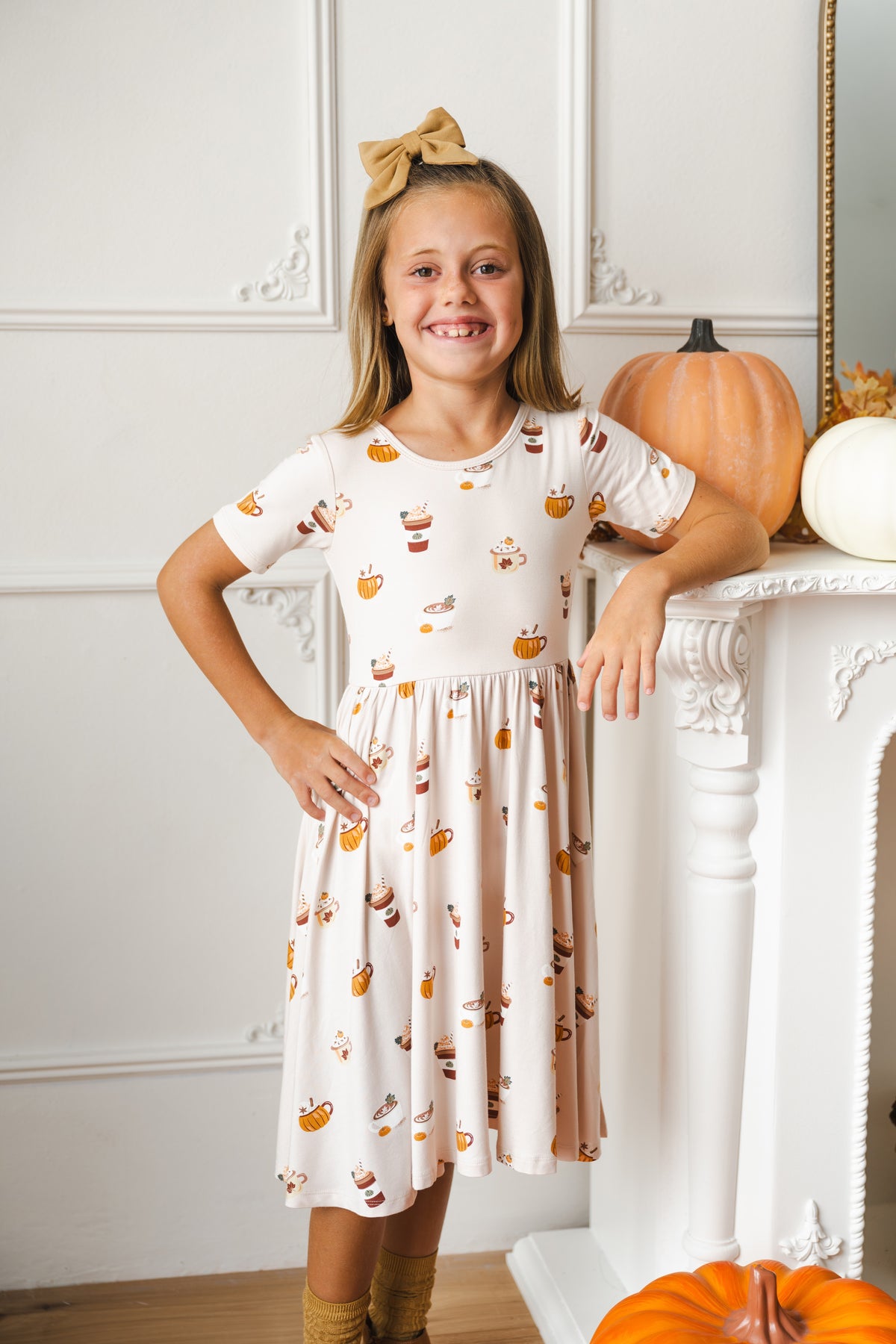 A young girl with a tan bow in her hair stands in a room with white paneled walls, leaning against a white fireplace mantel. She is wearing a cream-colored short-sleeve dress with a pumpkin and coffee cup pattern, paired with tan socks.