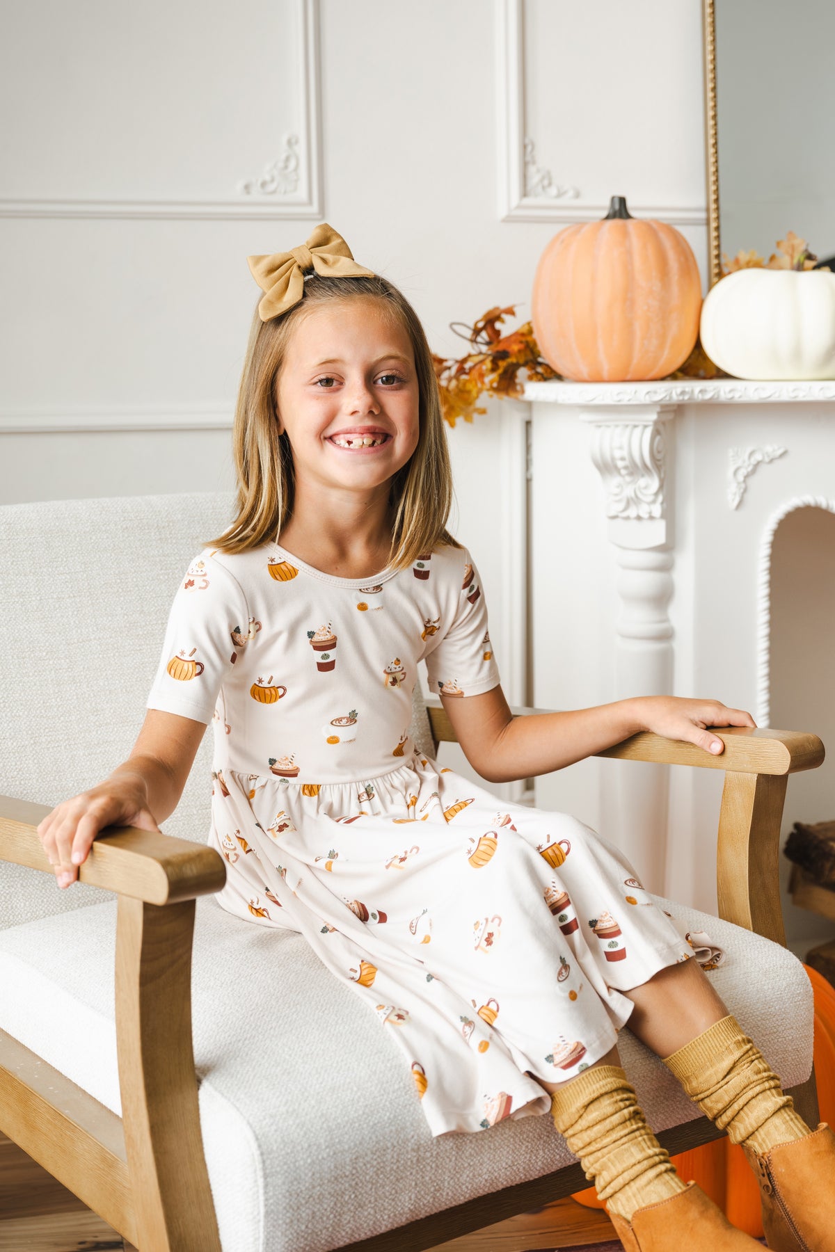 Girl wearing a pumpkin spice latte printed dress, tan bow and socks, sitting on a neutral chair