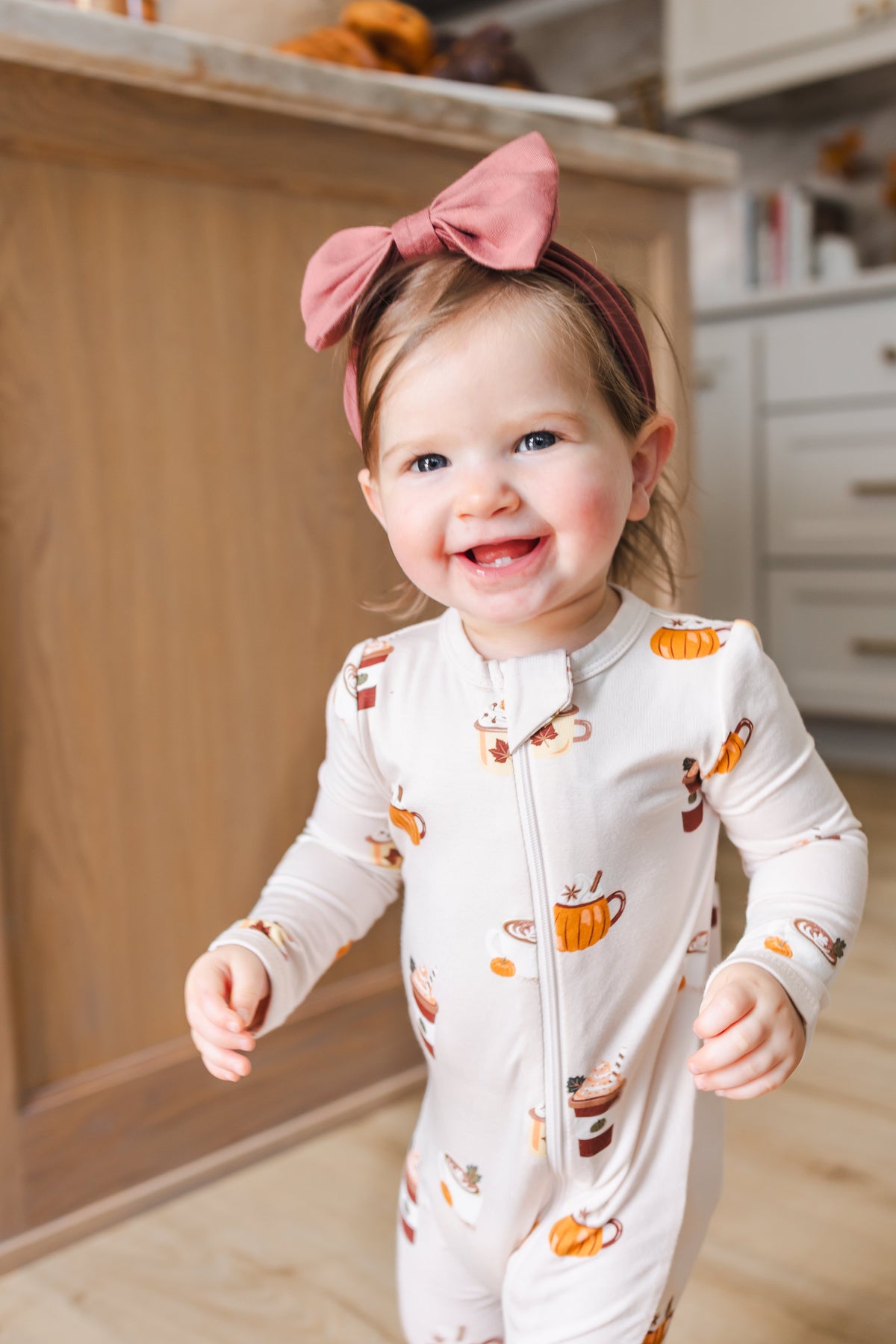 Close up of Toddler wearing Zippered Romper with Pumpkin Spice Latte print and pink knotted headband standing beside kitchen counter