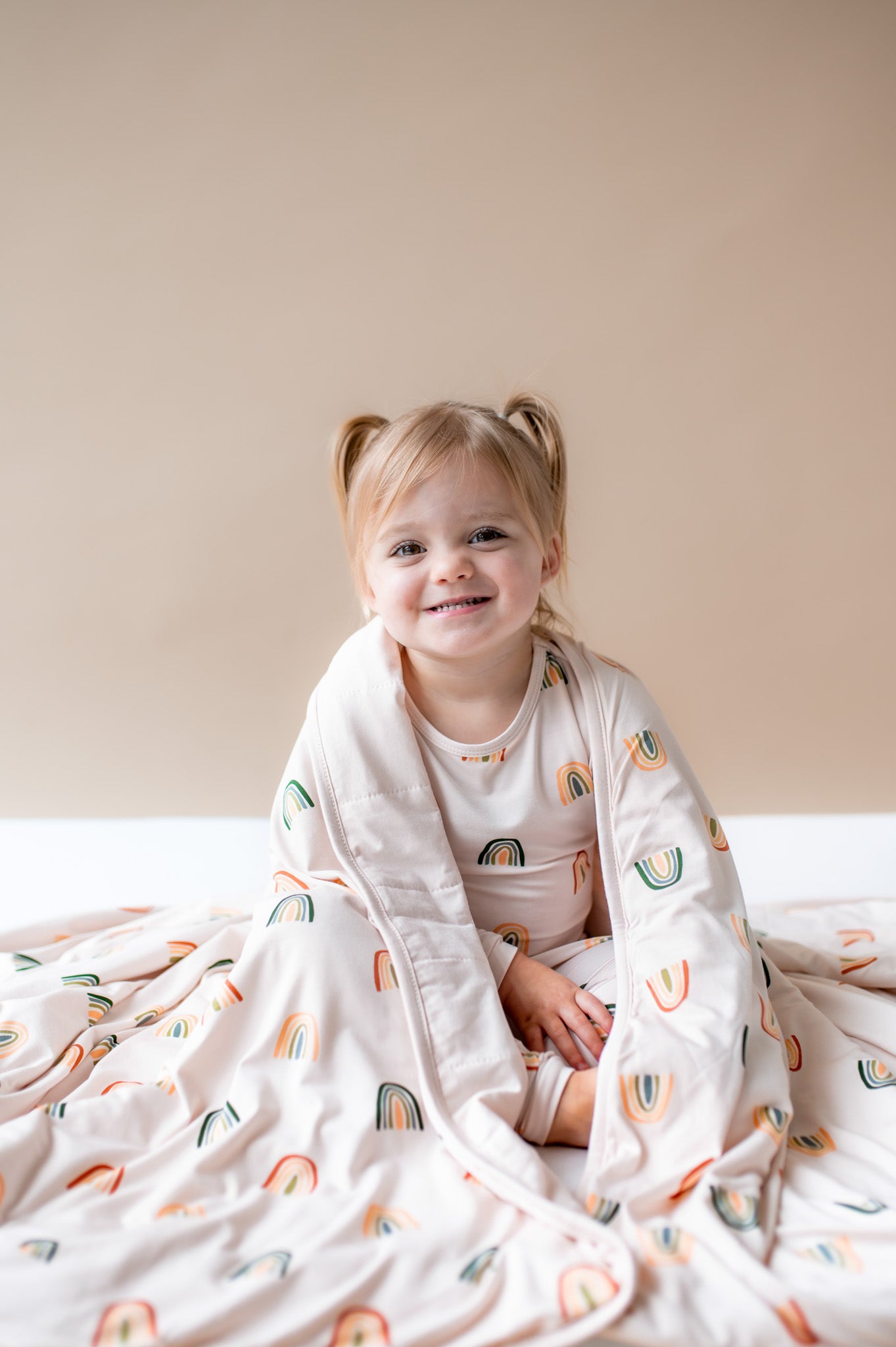 Girl wrapped in Toddler Blanket in Retro Rainbow 1.0 print sitting in a similar print long sleeve pajama set on a beige background