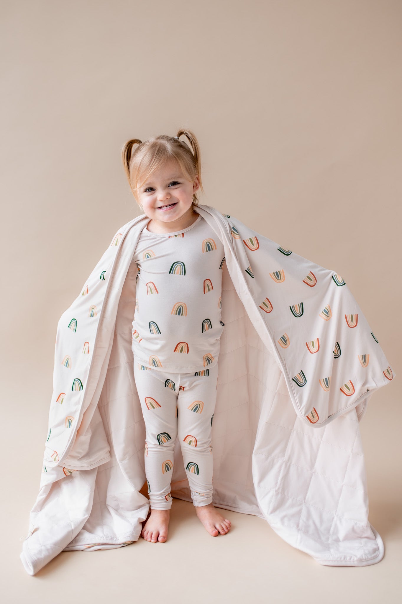 Girl holding Toddler Blanket in Retro Rainbow 1.0 print over her shoulders wearing a similar print long sleeve pajama set on a beige background