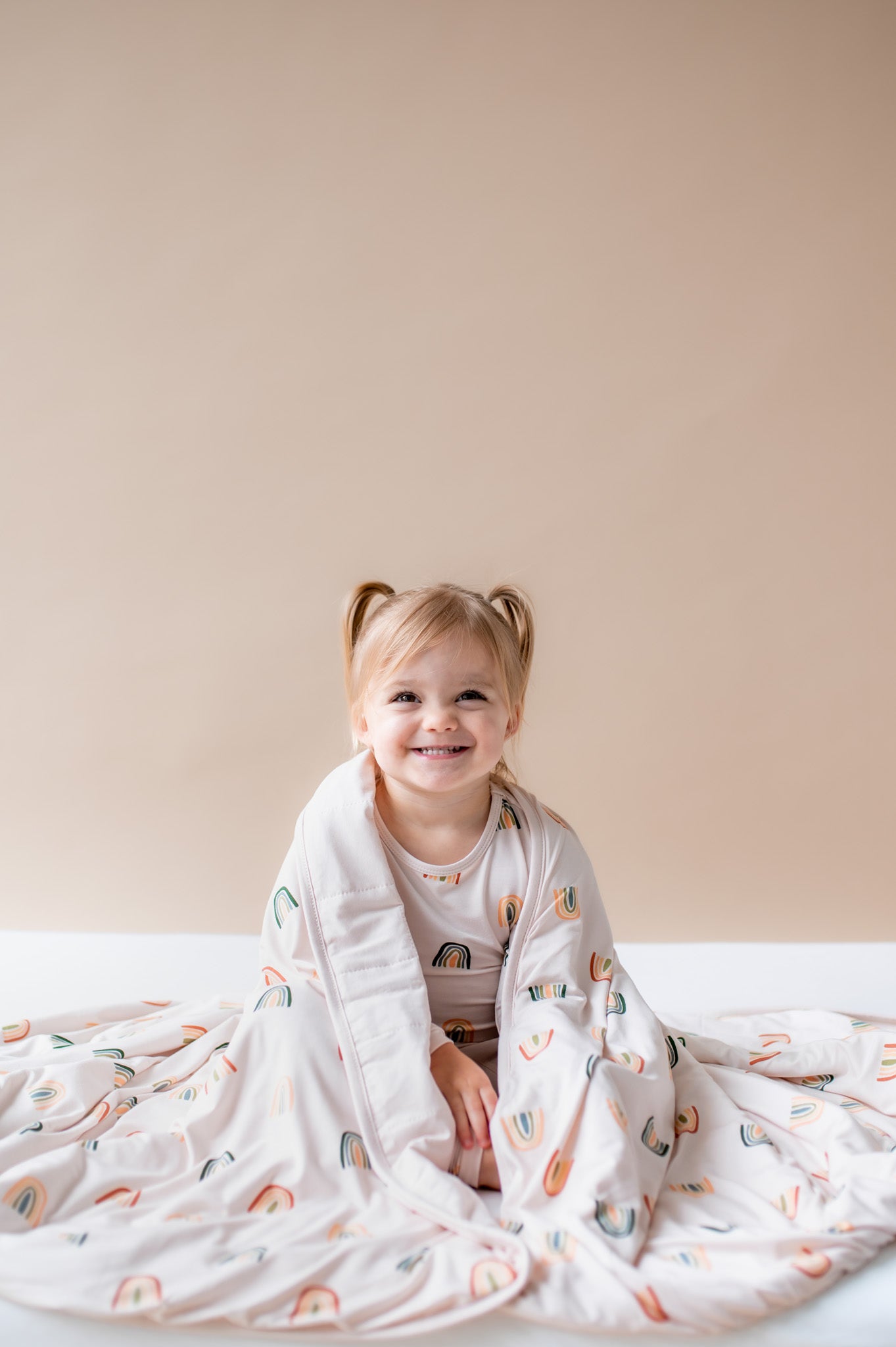 Girl sitting wrapped in Toddler Blanket in Retro Rainbow 1.0 print wearing a similar print long sleeve pajama set on a beige background