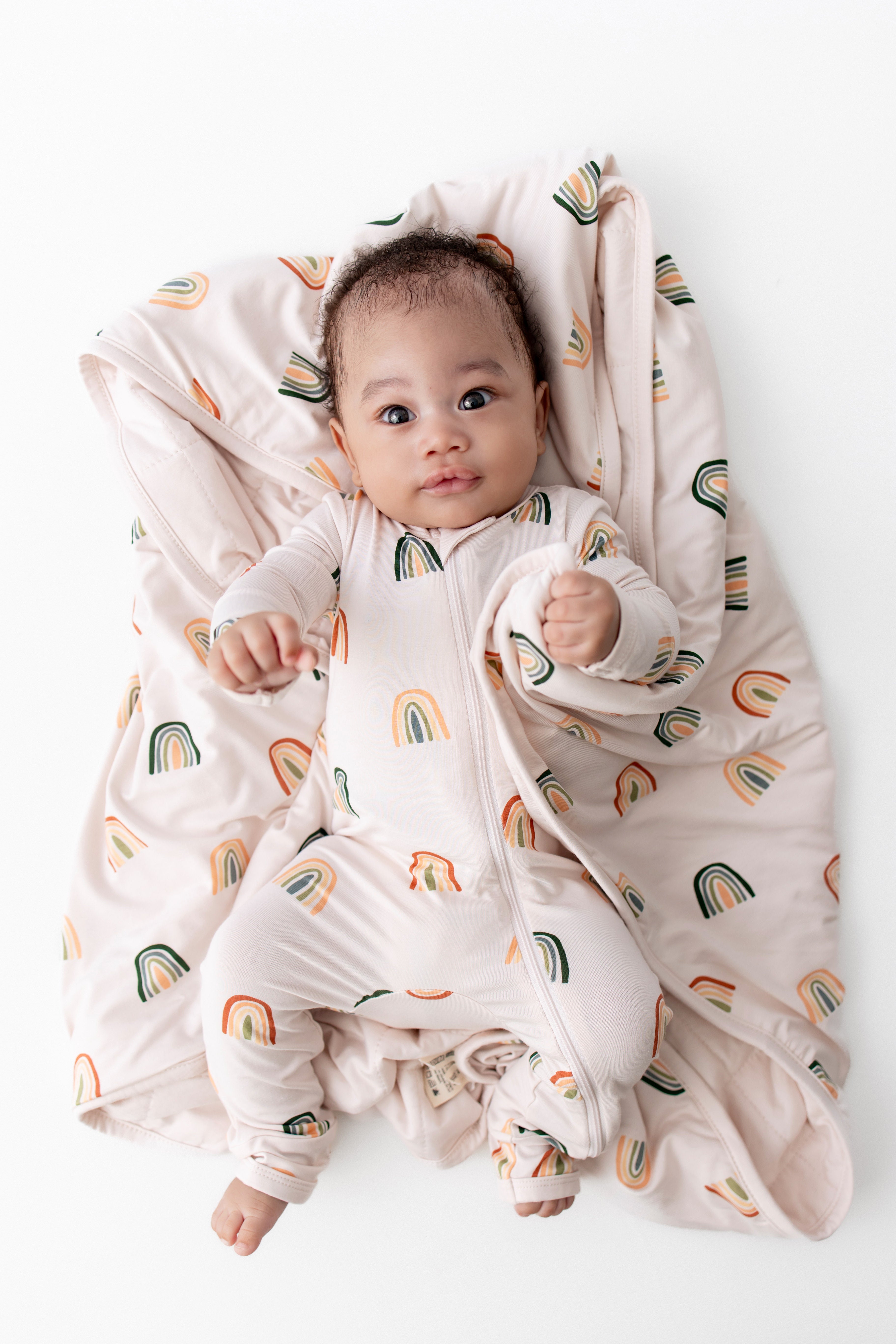 Baby wearing zippered romper with Retro Rainbow lying on Baby Blanket with Retro Rainbow print on a white background