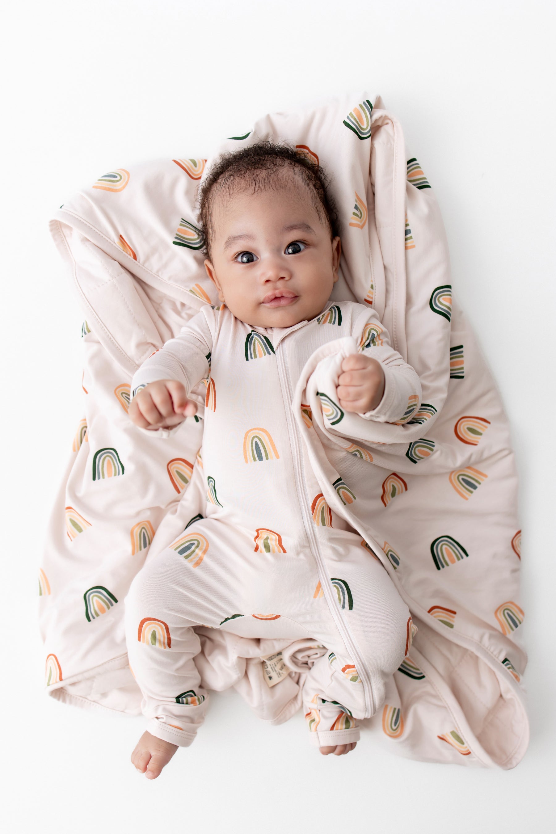 Baby wearing zippered romper with Retro Rainbow lying on Baby Blanket with Retro Rainbow print on a white background