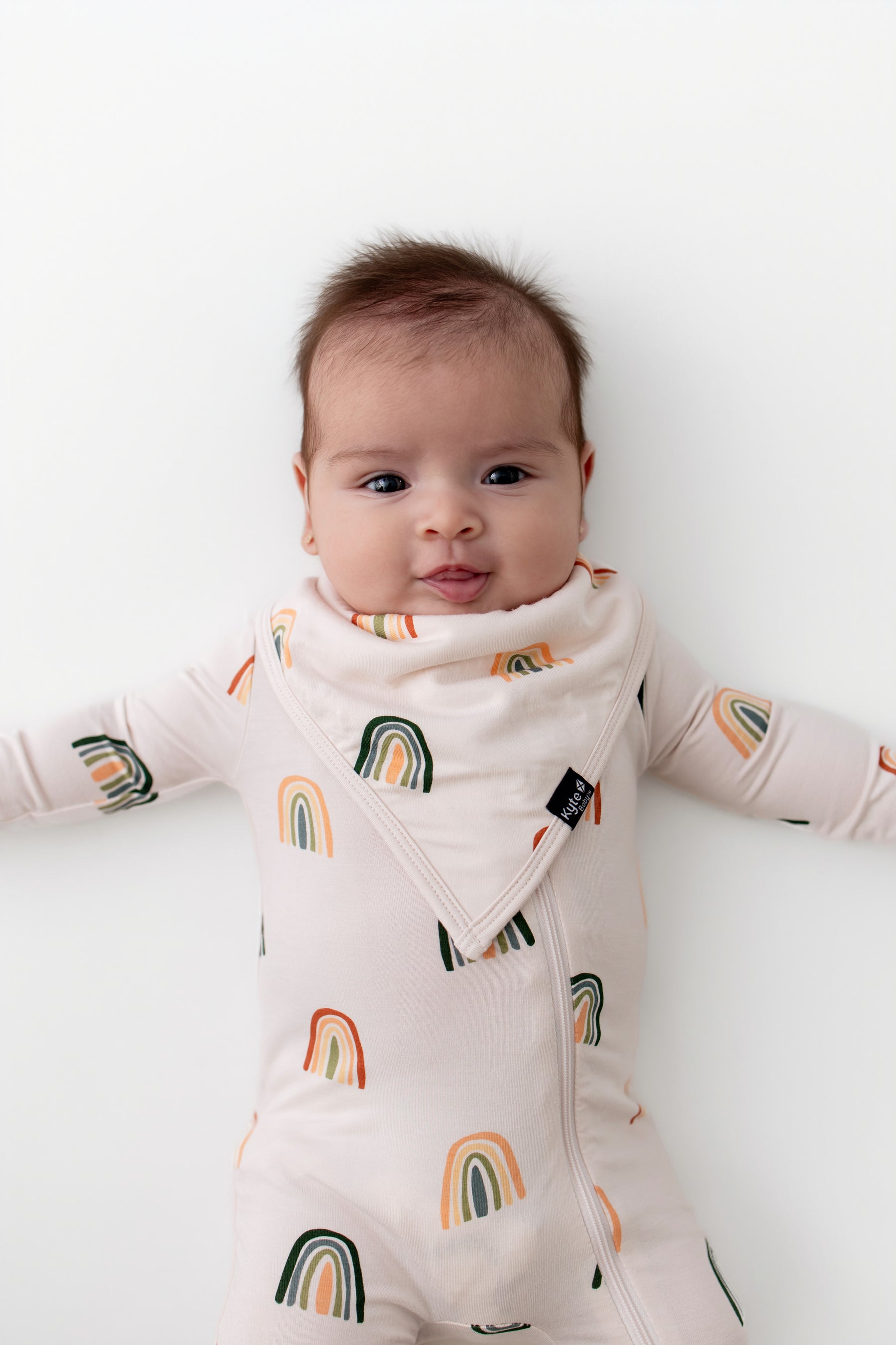Baby wearing Bib in Retro Rainbow print over a zippered romper with similar print on a white background