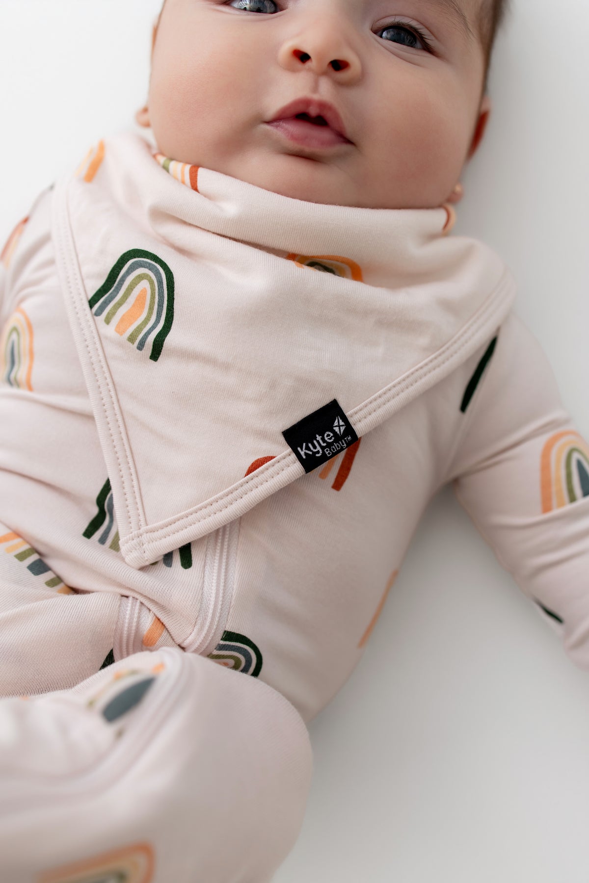 Baby wearing Bib in Retro Rainbow print over a zippered romper with similar print on a white background