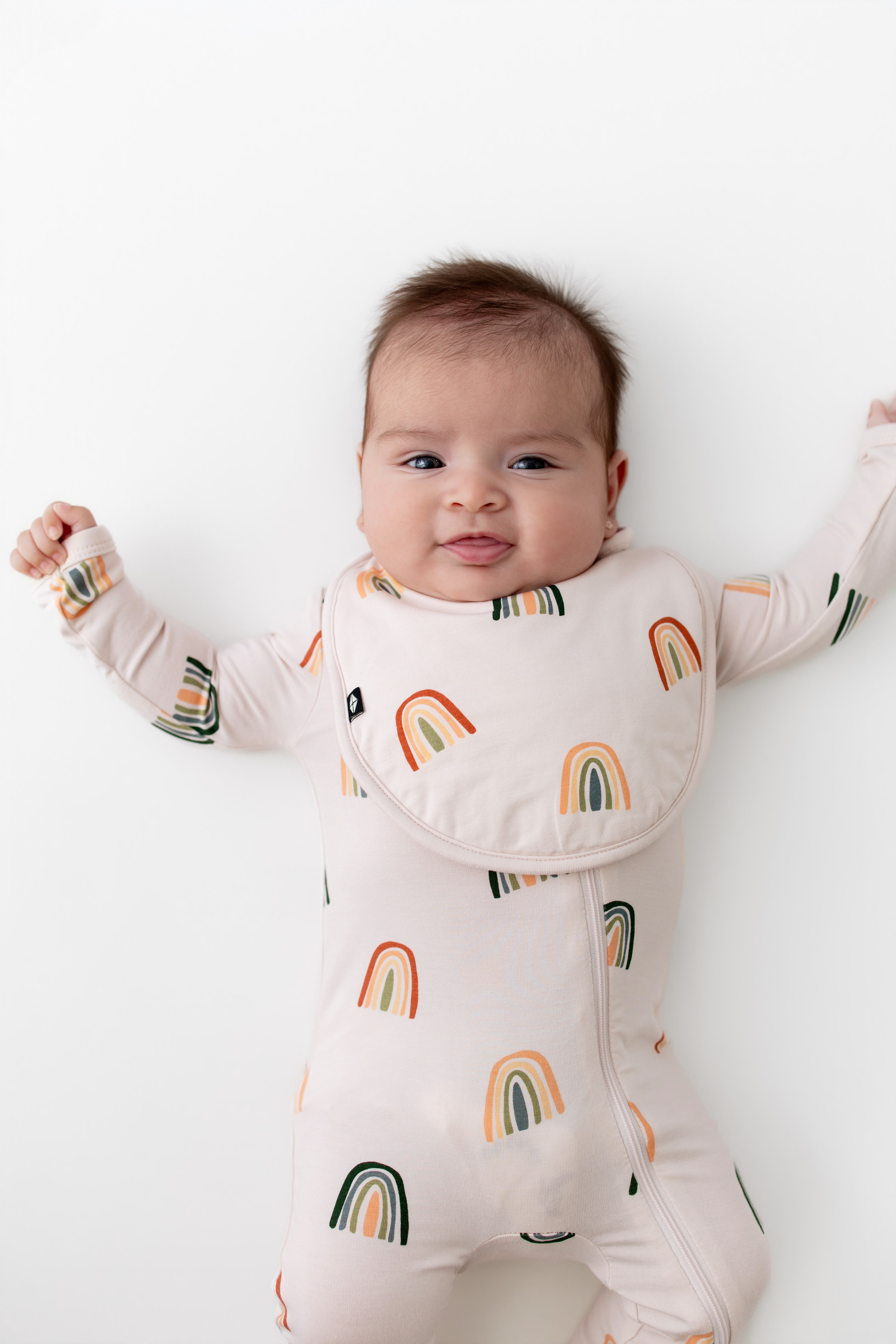 Baby smiling in Cushy Bib in Retro Rainbow print over a similar print zippered romper on a white background