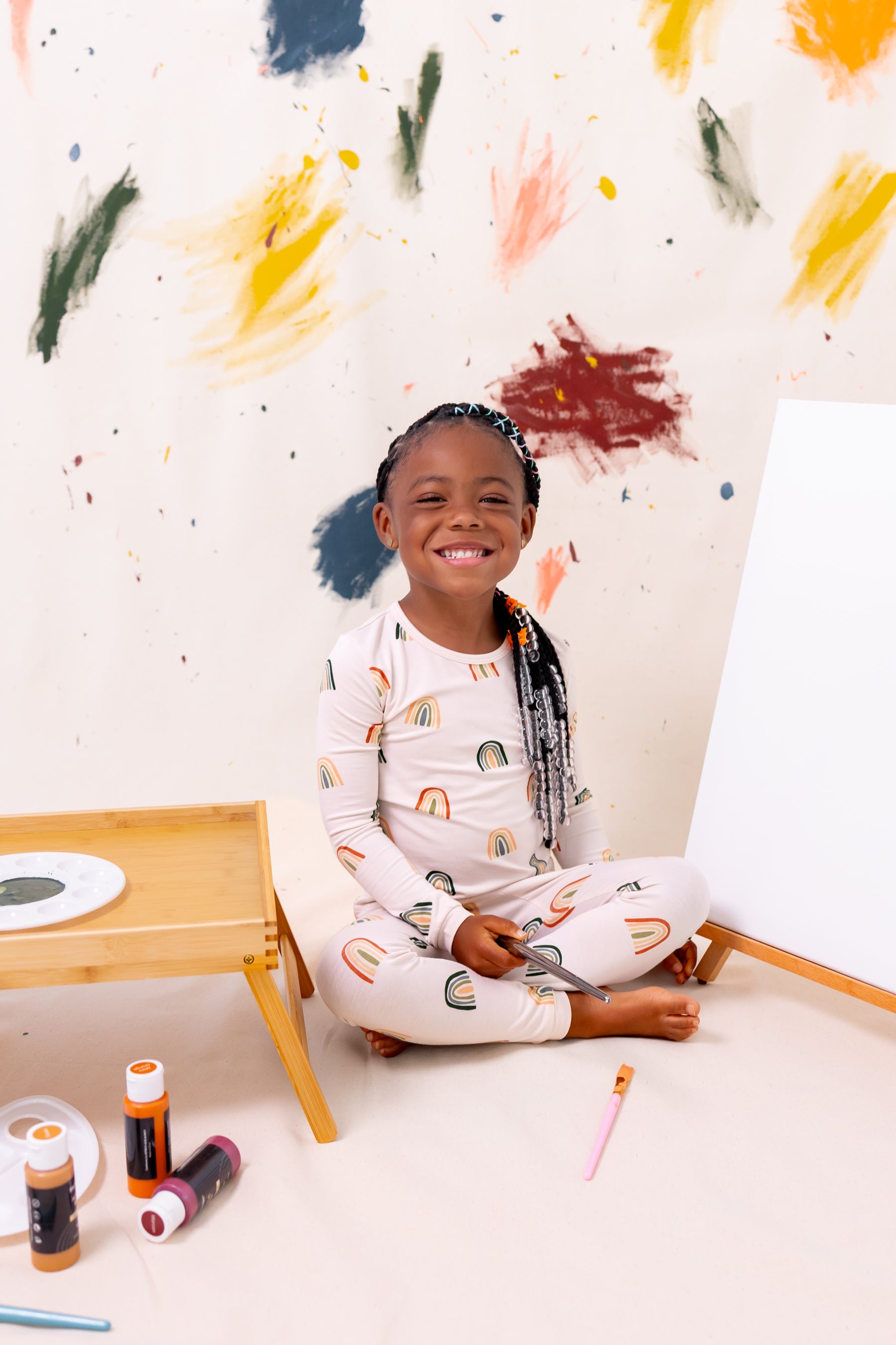 Girl sitting with paints wearing Long Sleeve Pajamas in Retro Rainbow print against a colorful background