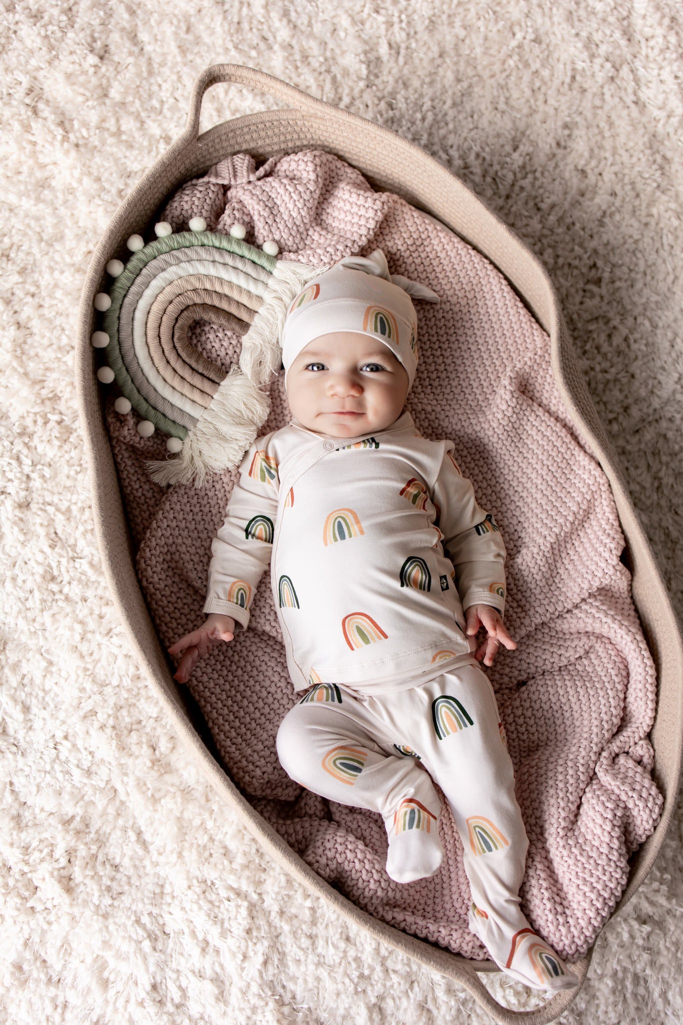 Baby lying in a basket over a pink blanket wearing Take Me Home Set with Cap in Retro Rainbow print with a retro rainbow prop