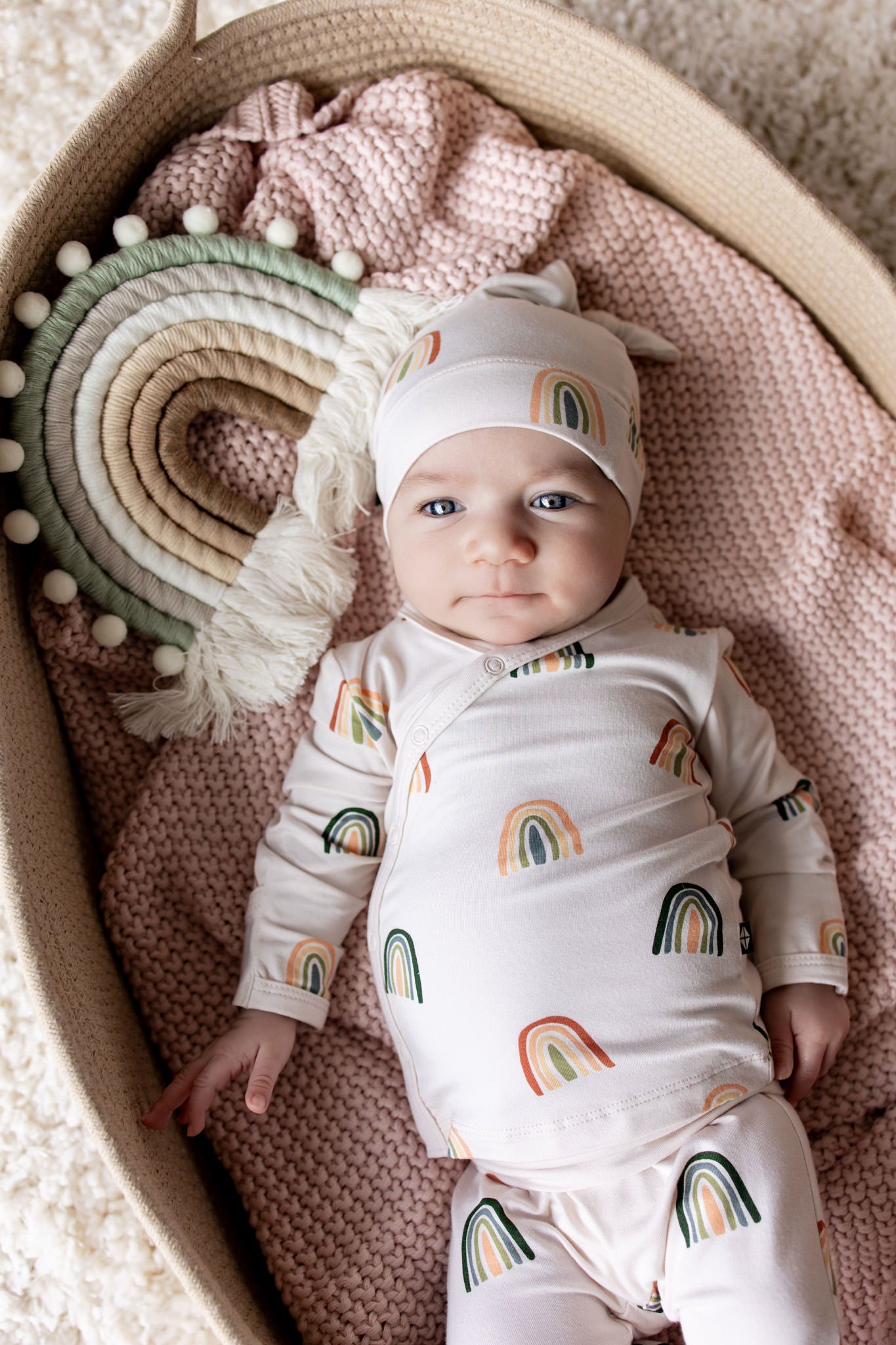Baby in Take Me Home Set with Cap in Retro Rainbow print lying in a basket over a pink blanket with a retro rainbow prop