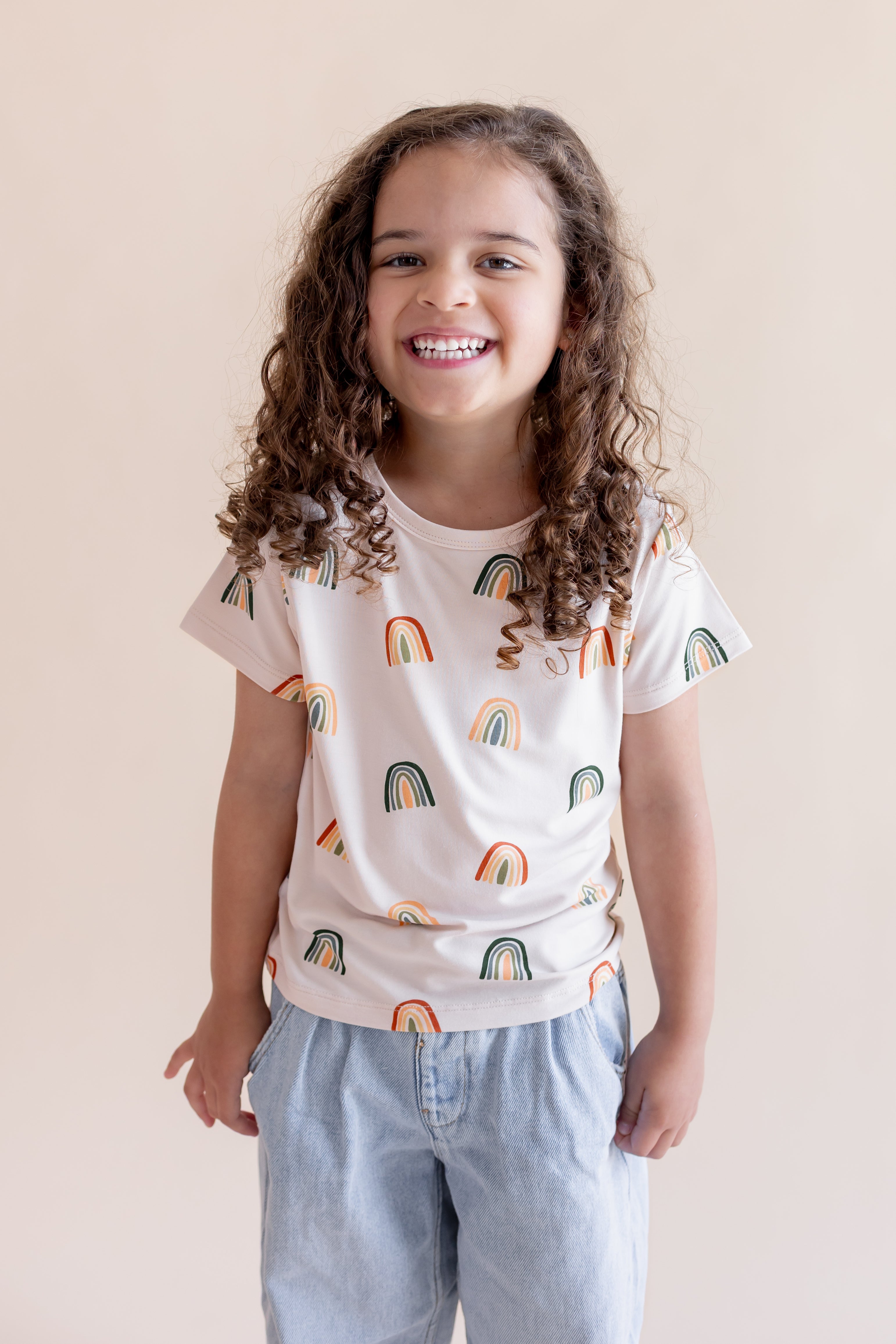 Girl posing in Toddler Basic Tee in Retro Rainbow print over blue pants on a white background