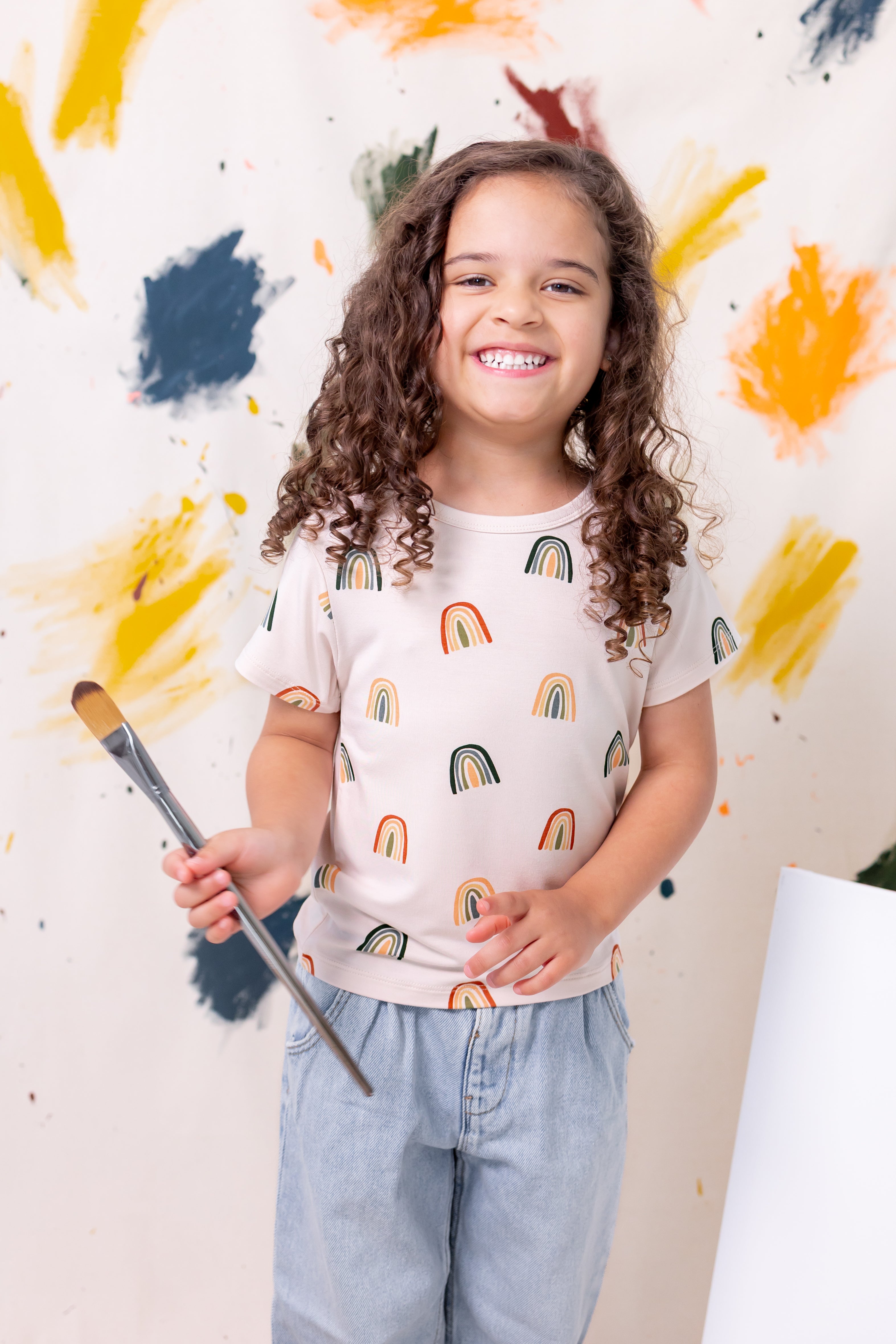 Girl wearing Toddler Basic Tee in Retro Rainbow print over blue pants holding a paint brush in front of a colorful background