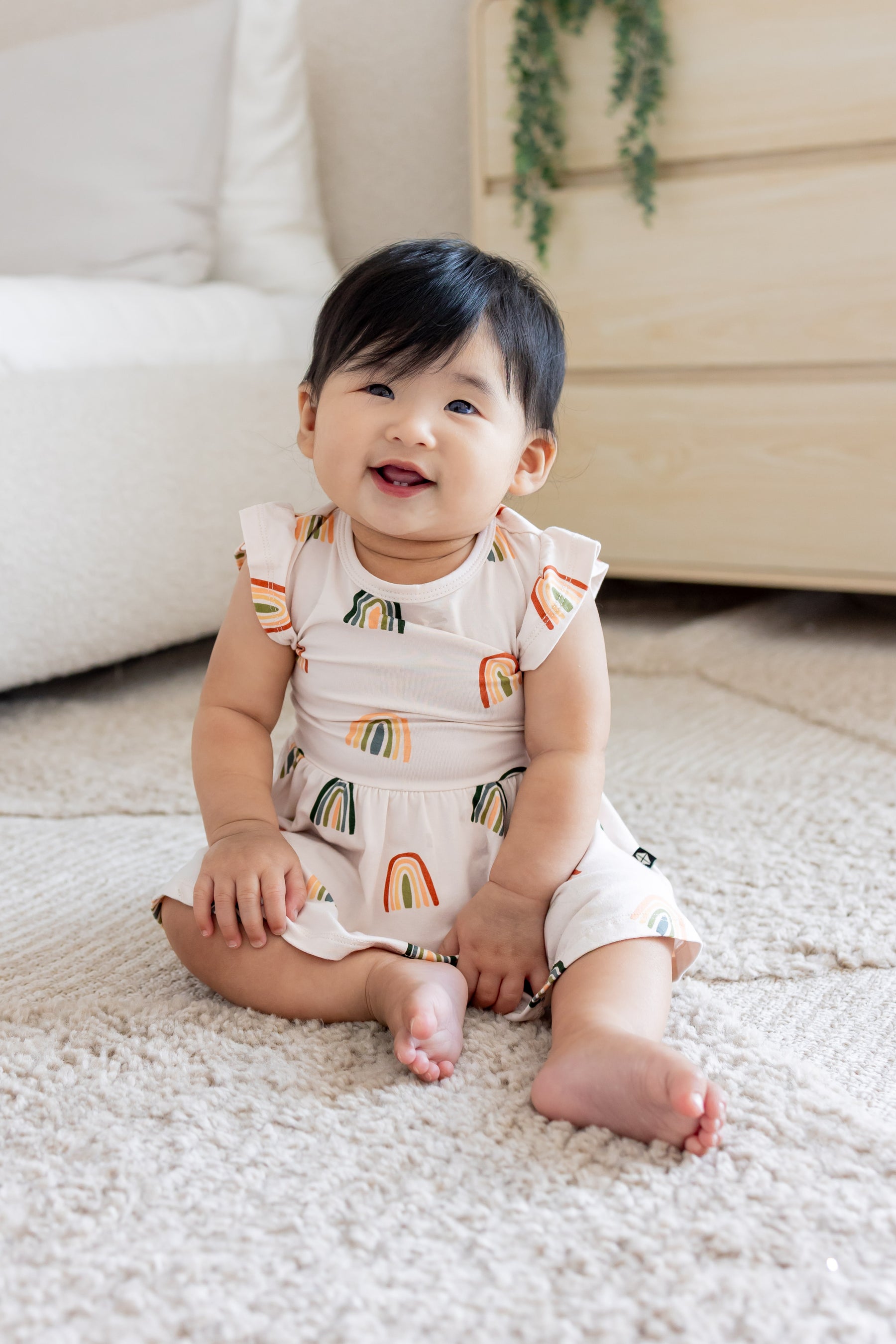Baby sitting on a white carpet wearing Twirl Bodysuit Dress in Retro Rainbow print 
