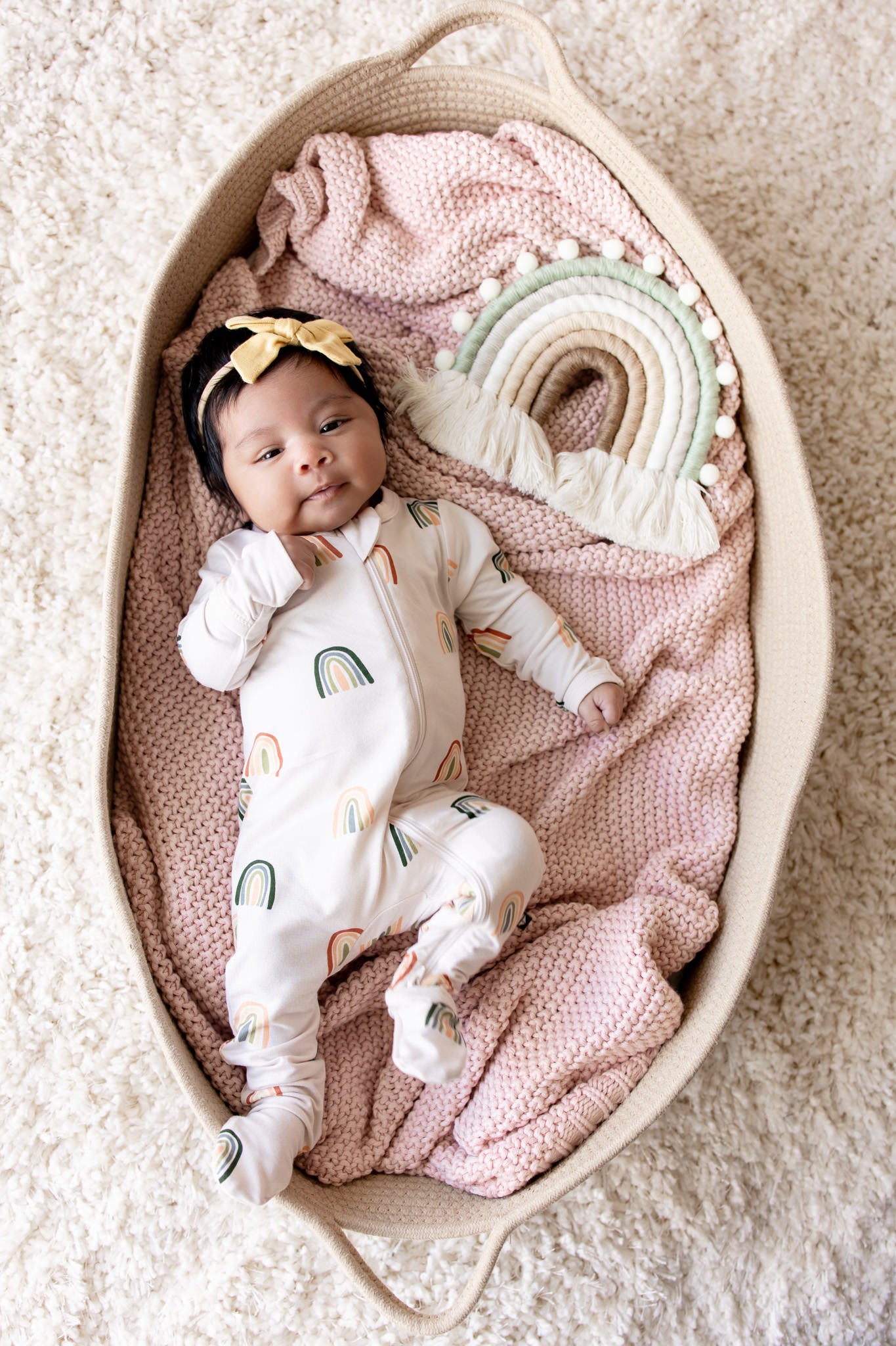 Baby lying in a basket over a pink blanket wearing Zippered Footie in Retro Rainbow print with a retro rainbow prop