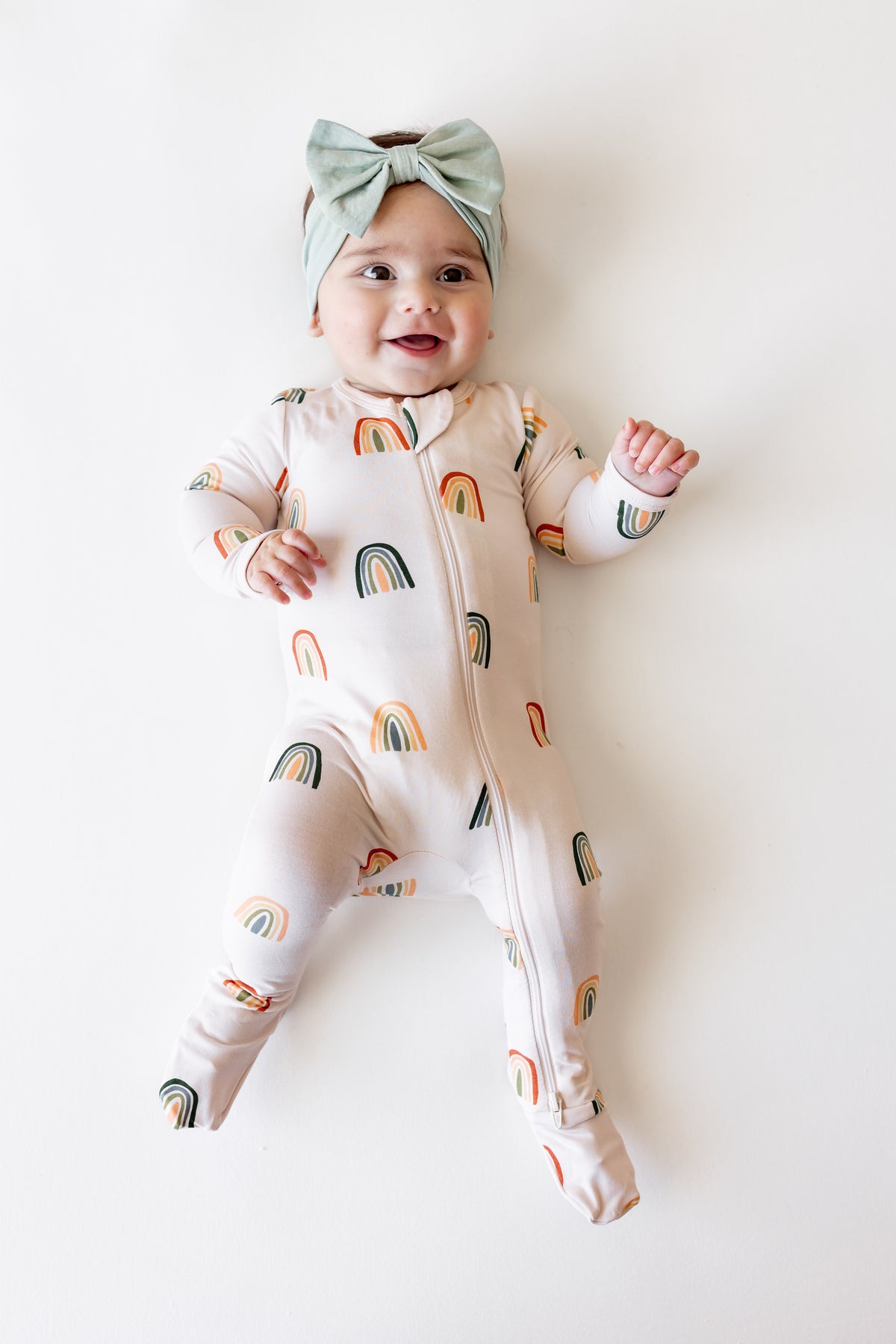Baby wearing Zippered Footie in Retro Rainbow print and a blue bow on head on a white background
