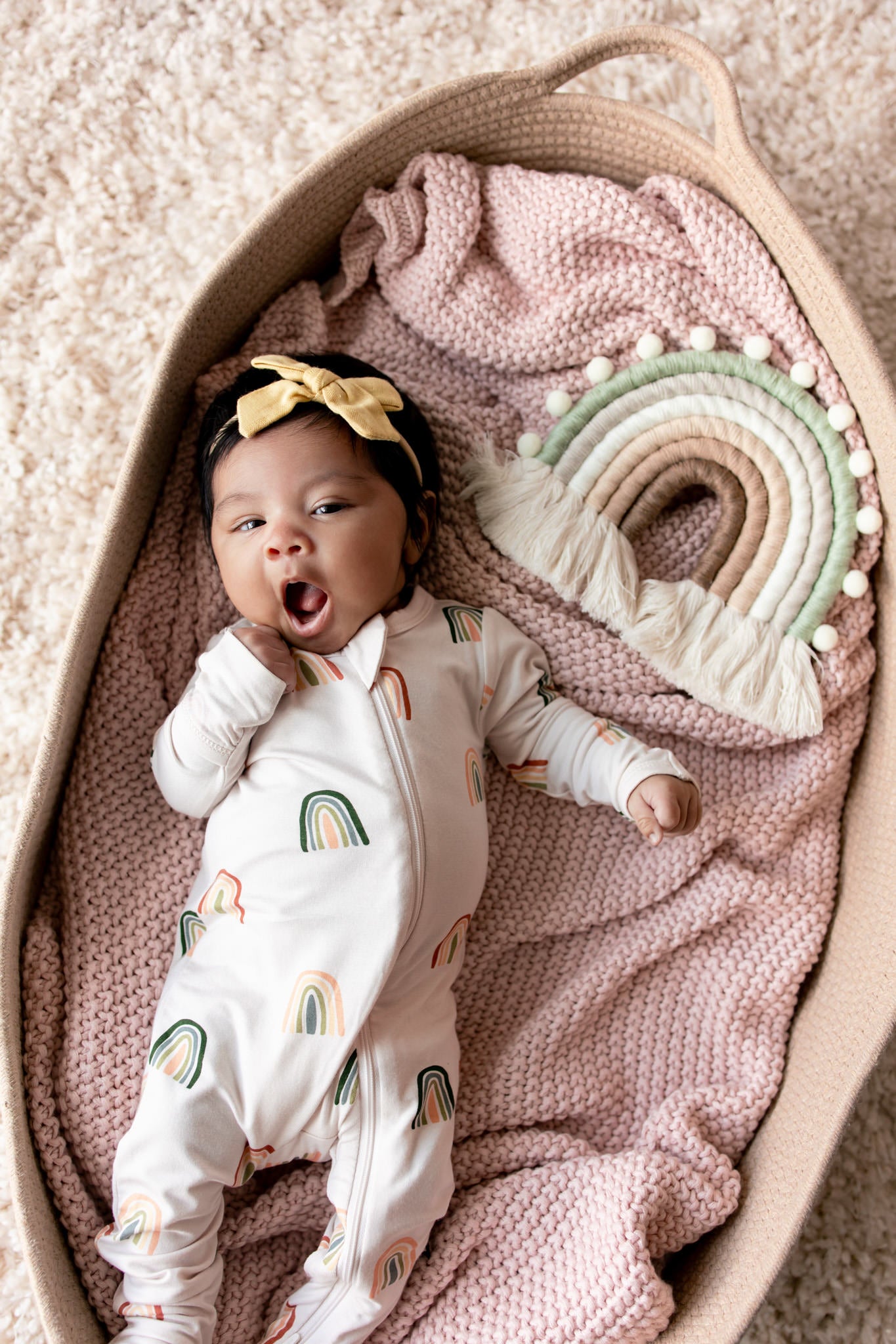 Baby yawning in a basket over a pink blanket wearing Zippered Footie in Retro Rainbow print with a retro rainbow prop