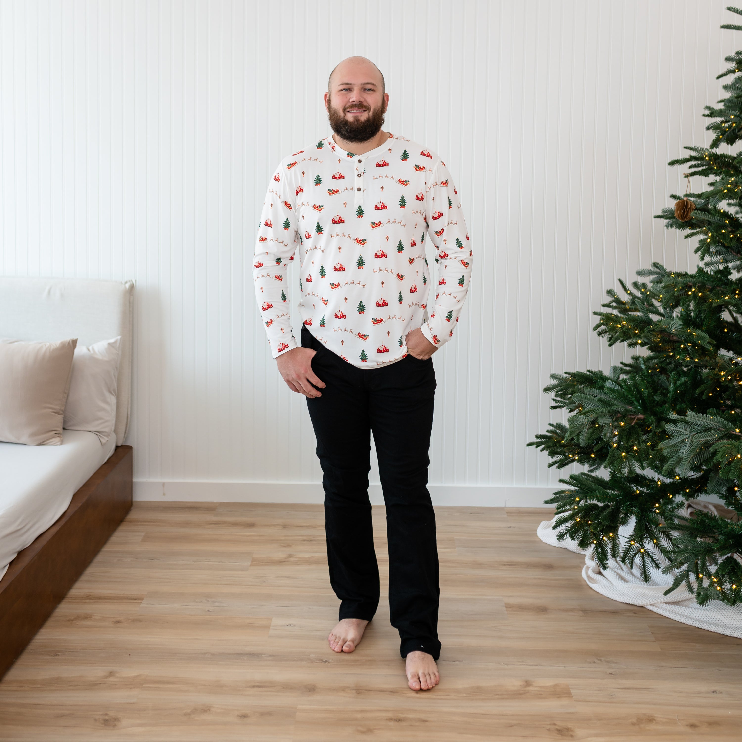 Male model standing beside a christmas tree wearing the Men's Long Sleeve Henley Top in Santa Sleigh with his hands in his black jeans pocket