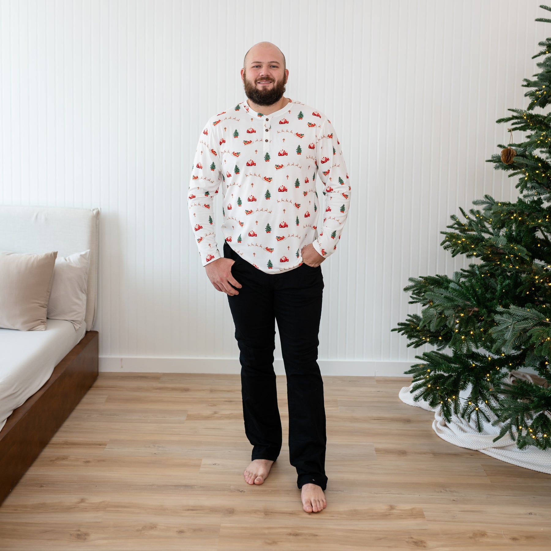 Male model standing beside a christmas tree wearing the Men's Long Sleeve Henley Top in Santa Sleigh with his hands in his black jeans pocket