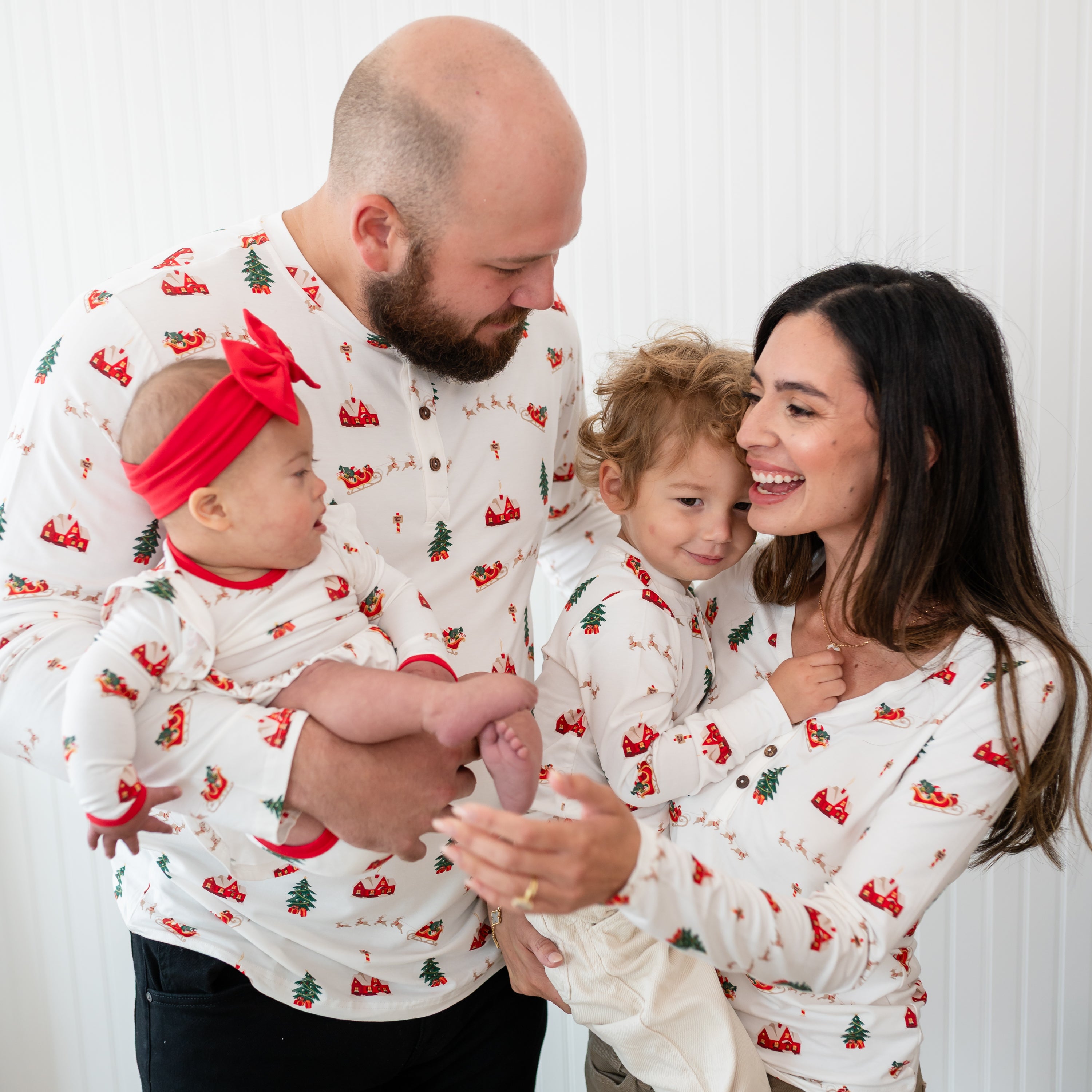 Family of four matching in Santa Sleigh. Father, Mother and Son are all wearing a Henley tee with the daughter wearing a long sleeve twirl bodysuit dress