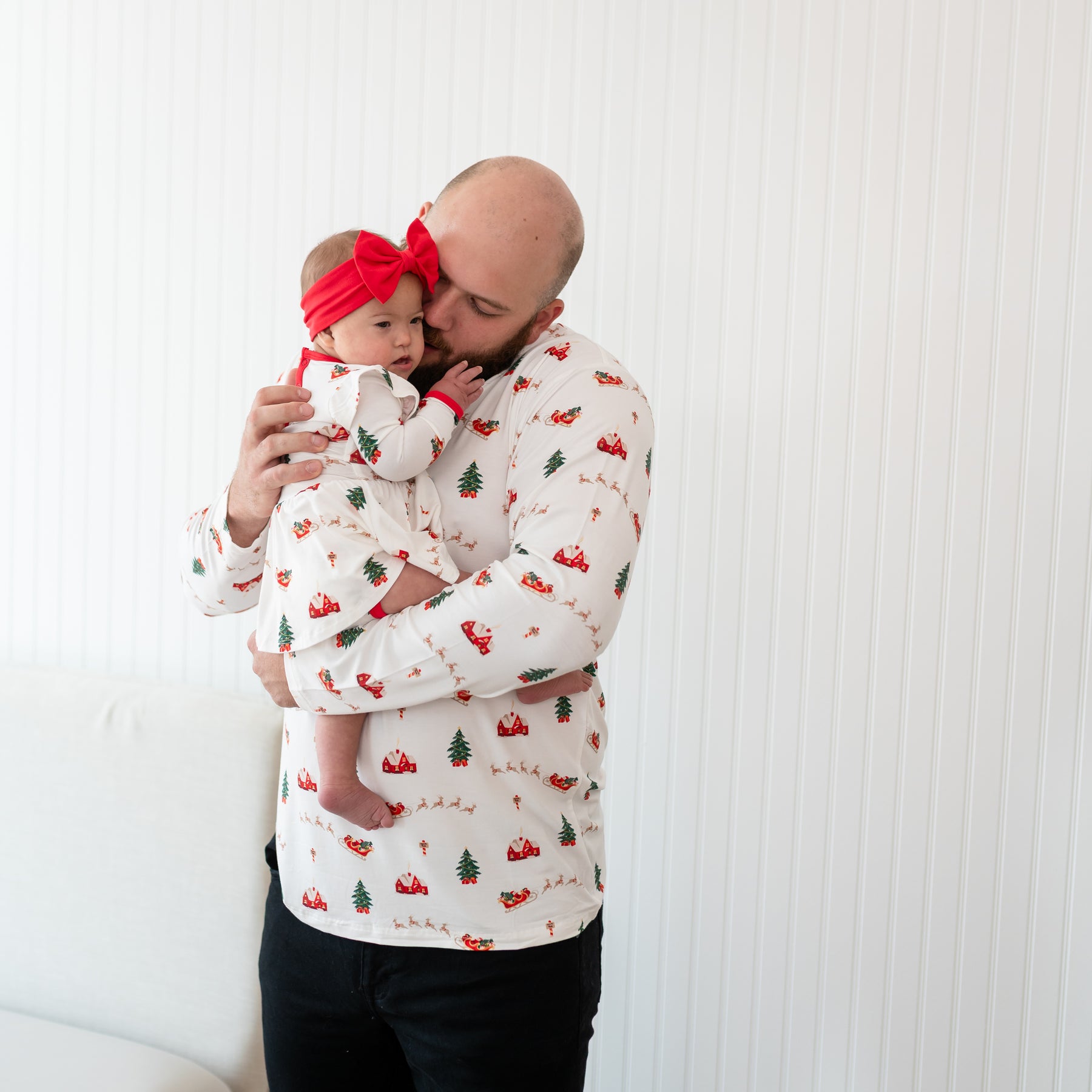 Father wearing the Men's Henley Top in Santa Sleigh holding his infant daughter wearing the Long Sleeve Twirl Bodysuit Dress in Santa Sleigh and Cardinal bow headband
