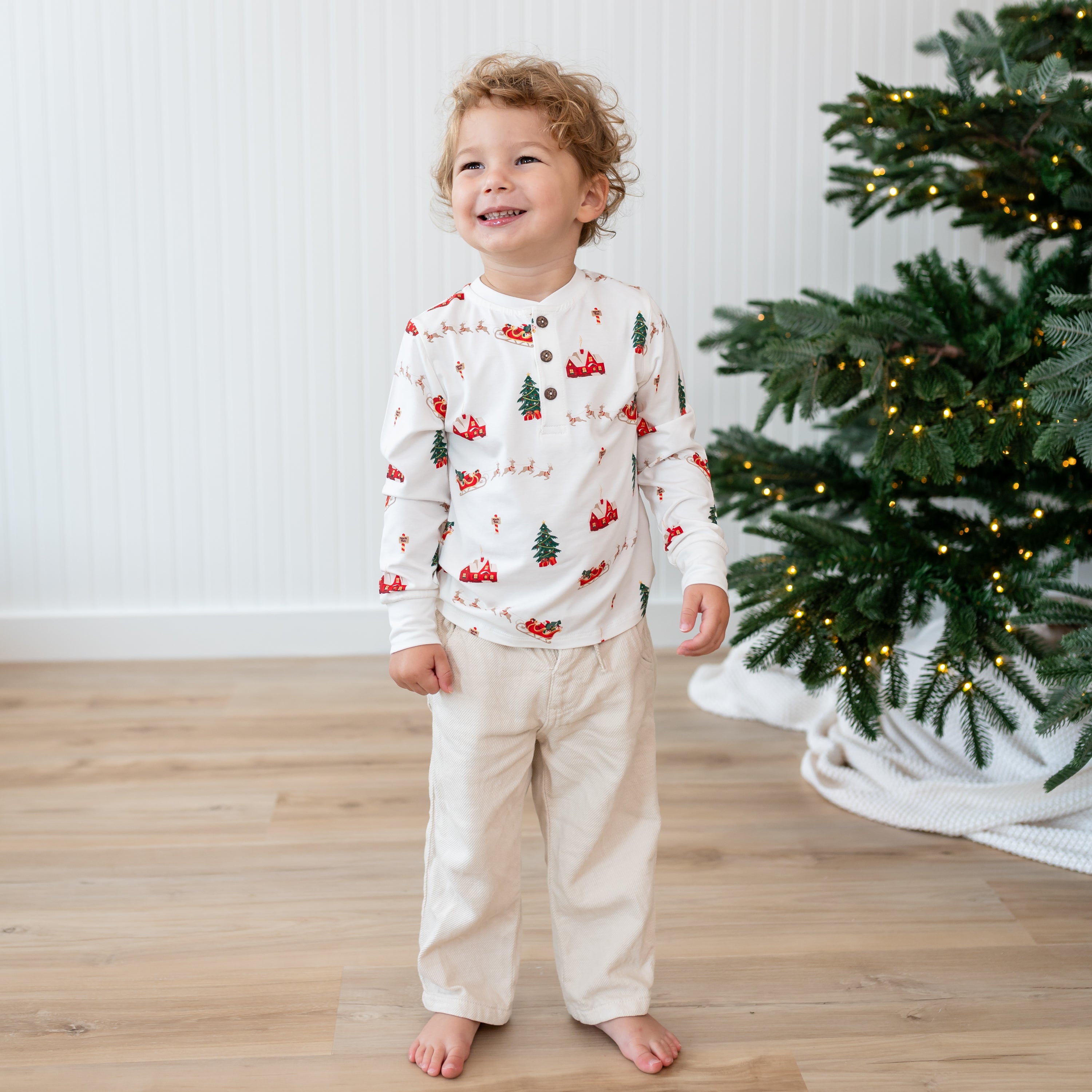 Smiling boy standing in front of a christmas tree wearing the Long Sleeve Toddler Henley Tee in Santa Sleigh with cream colored pants