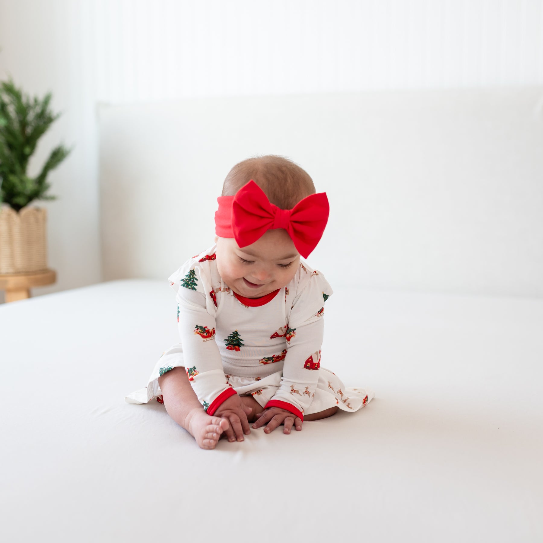 Infant sitting on a bed wearing the Bow Headband in Cardinal and Long sleeve twirl bodysuit dress in Santa Sleigh