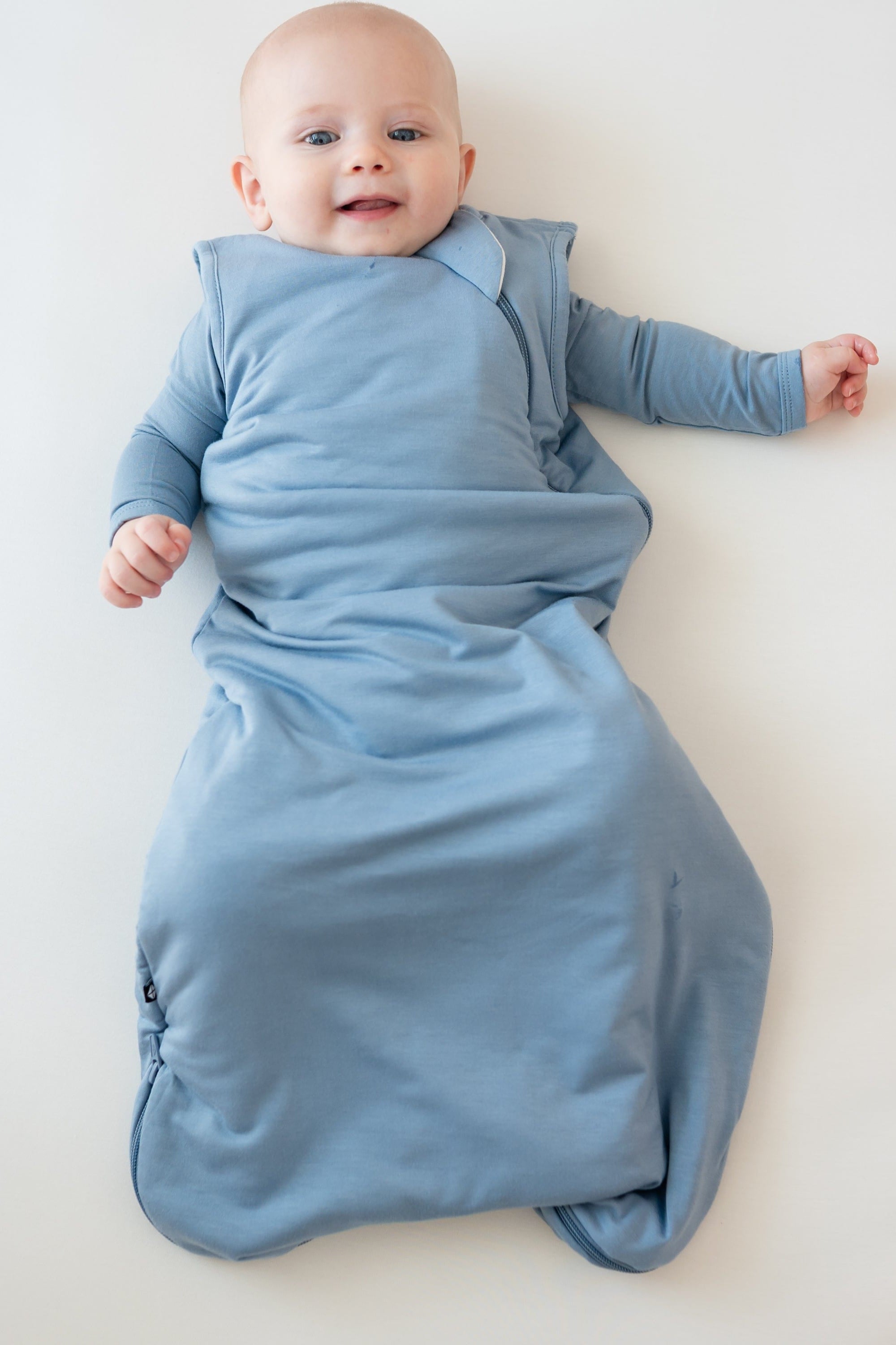 Baby laying in crib wearing Kyte Baby Sleep Bag in Slate 1.0 with matching bodysuit underneath